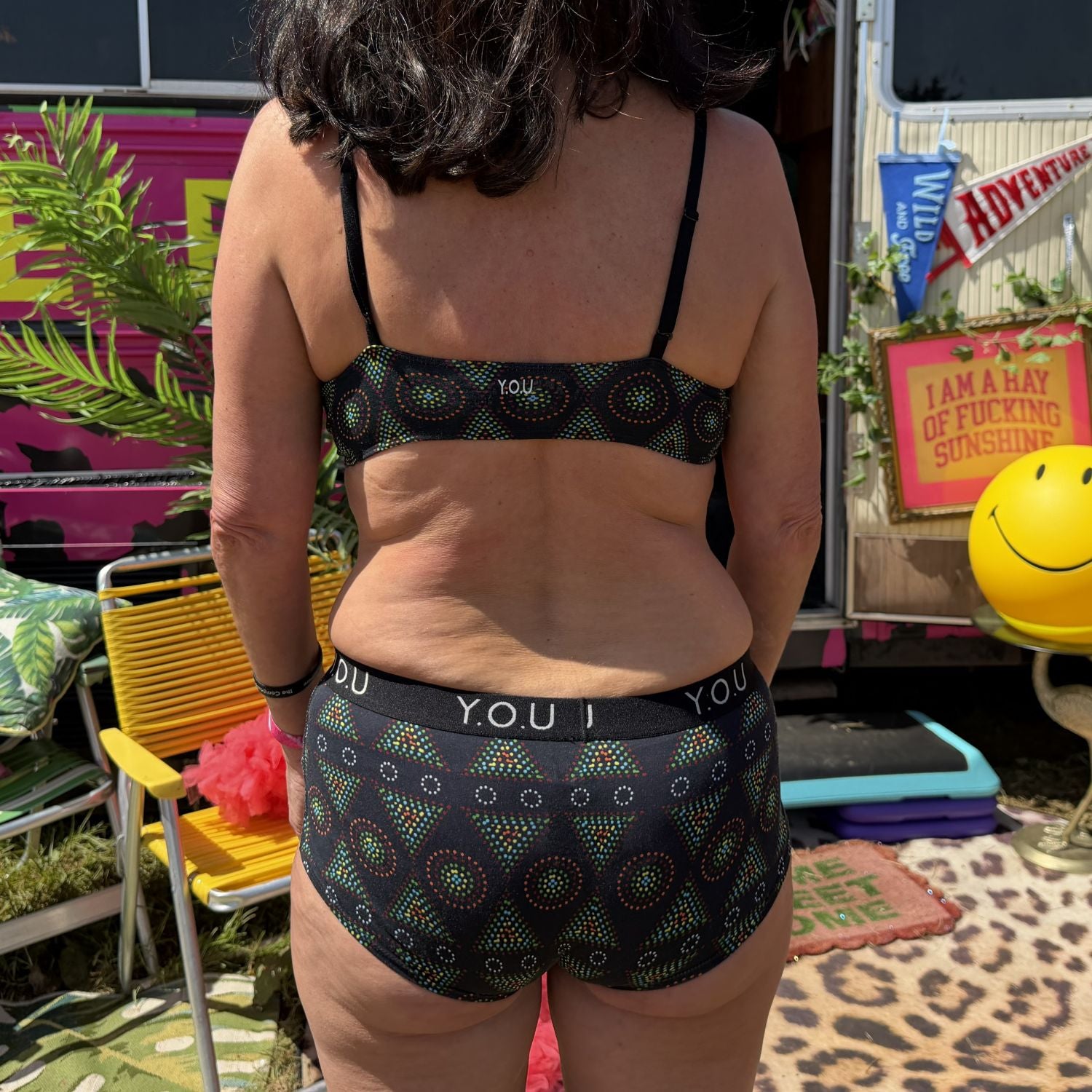 A photo of a model wearing the YOU Underwear bralette and boyshorts in black mara. The photo is zoomed in so we can see from the model's shoulder to their mid thigh. They are facing away from the camera, with their hands at their sides. The background is busy and colourful with greenery, flags and camping chairs.