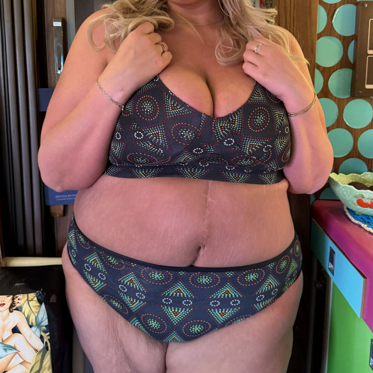 A photo of a model wearing the YOU Underwear non wired bra and mid rise bikini knickers in the black mara print. The photo is zoomed in so we can only see from the model's shoulders to their upper-thigh. They are holding the straps of their bra with both their hands. 