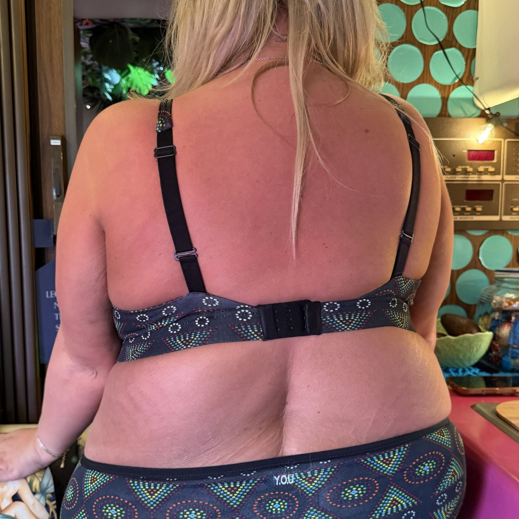 A photo of a model wearing the YOU Underwear black mara non-wired bra and matching knickers. The photo is zoomed in so we can see from the model's shoulder's to their hips and they are facing away from the camera with their hands at their sides. Behind them is a wooden wall with bright blue polka dots on.