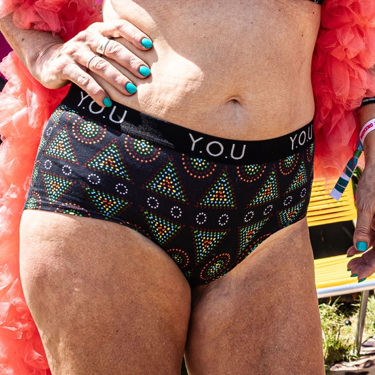 A photo of a model wearing the YOU Underwear branded boyshorts in the black mara print. The photo is zoomed in so we can see from the model's stomach to their upper-thigh. She has her right arm on her hip and the other at her side. She is wearing a pink feather bower which we can see in the photo.