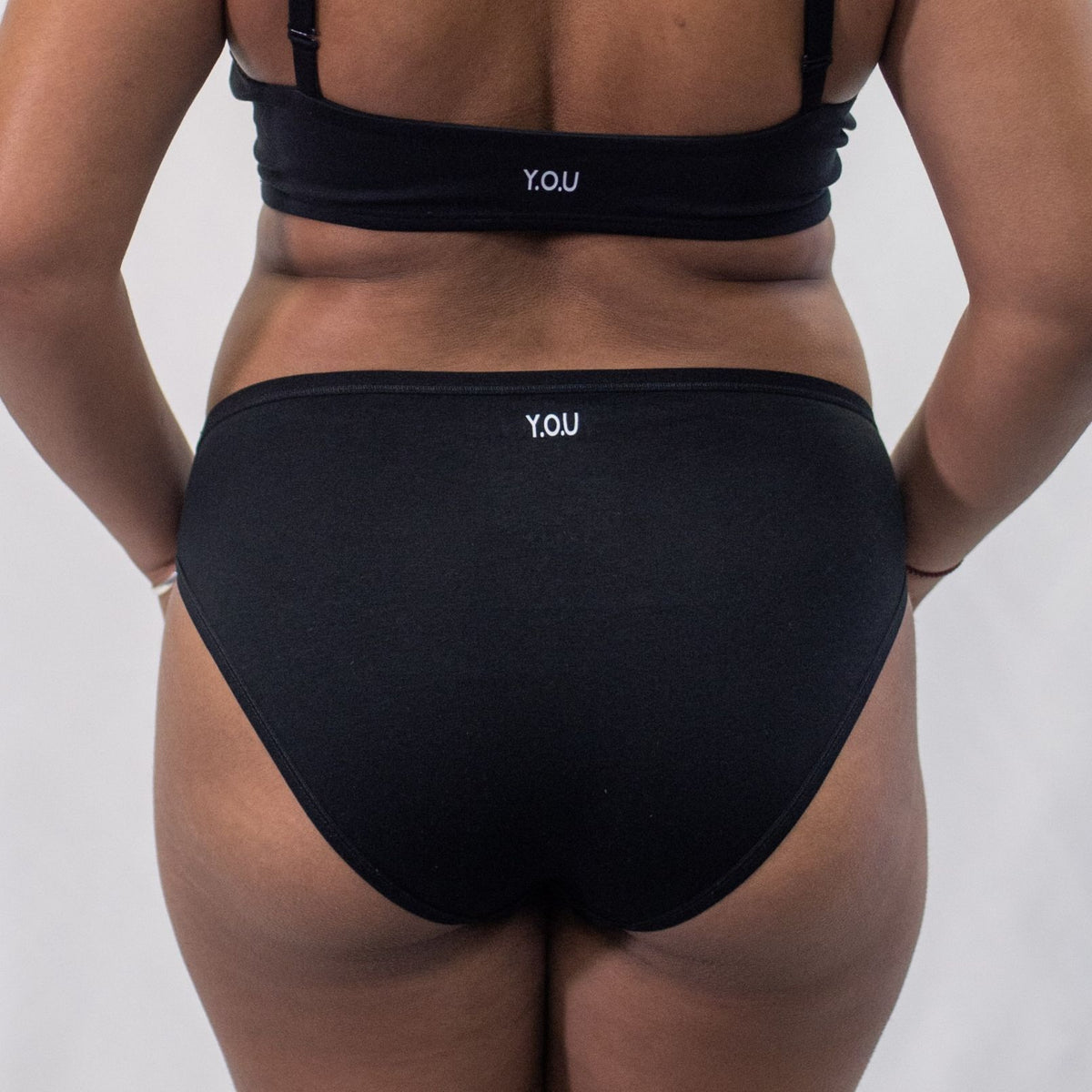 A photo of a model wearing the YOU Underwear midrise bikini knickers in black. The model is standing facing the camera with their hands in front of them. The photo is zoomed in so we can see from the model's stomach to their upper thigh. The background is white.