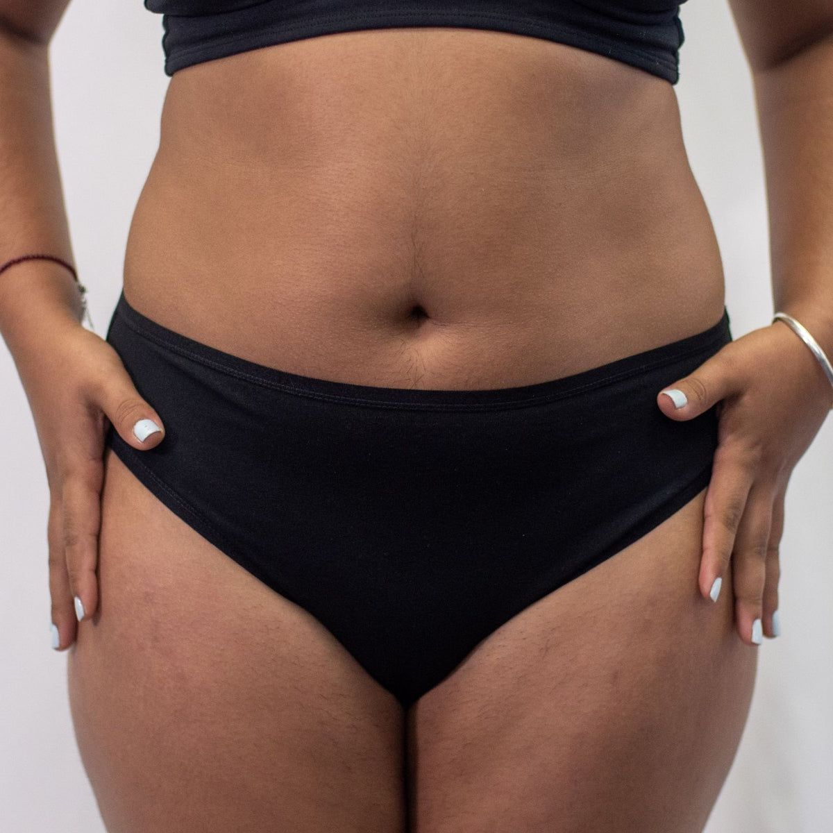A photo of a model wearing the YOU Underwear midrise bikini knickers in black. The model is standing facing the camera with their hands flat on their hips. The photo is zoomed in so we can see from the model's stomach to their upper thigh. The background is white.