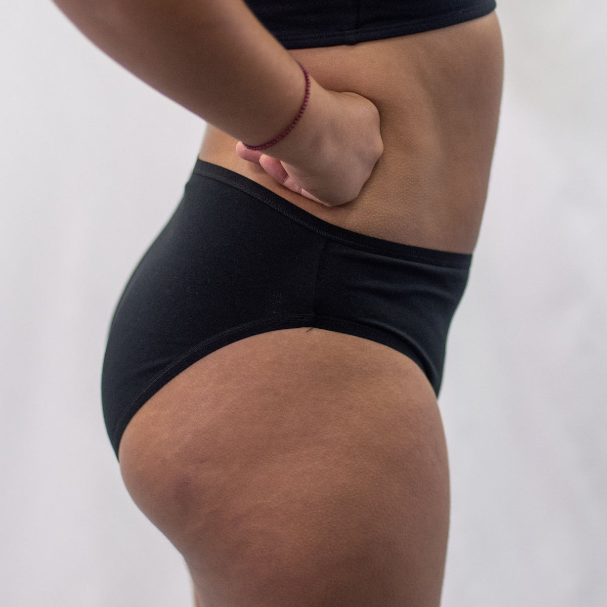 A photo of a model wearing the YOU Underwear midrise bikini knickers in black. The model is standing side on to the camera with their hands on their hips. The photo is zoomed in so we can see from the model's stomach to their upper thigh. The background is white.