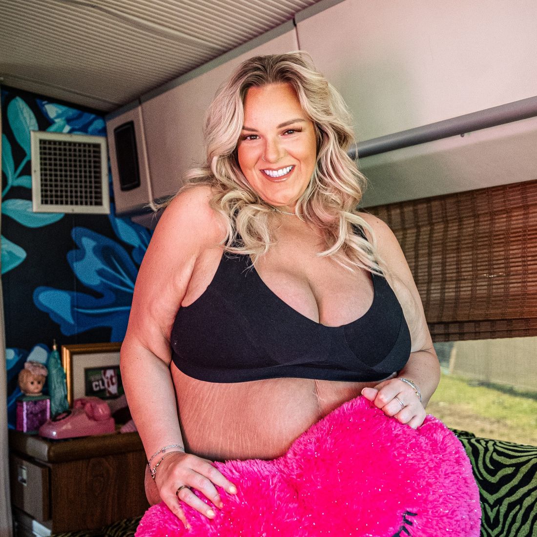 A photo of a model wearing the YOU Underwear non-wired bra in black. The photo is zoomed in so we can only see from the model's head to their stomach and they are holding a hot pink heart shaped cushion over her stomach. She is face on to the camera, smiling. 