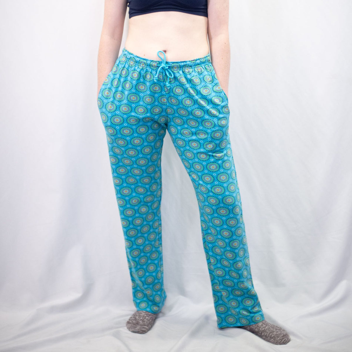 A photo of a model wearing the YOU Underwear pyjama bottoms in the vibrant blue mara print. The model is standing facing the camera with their hands in their pockets. They are wearing grey fluffy socks and we can see from the model's stomach to the floor. There is a whie background.