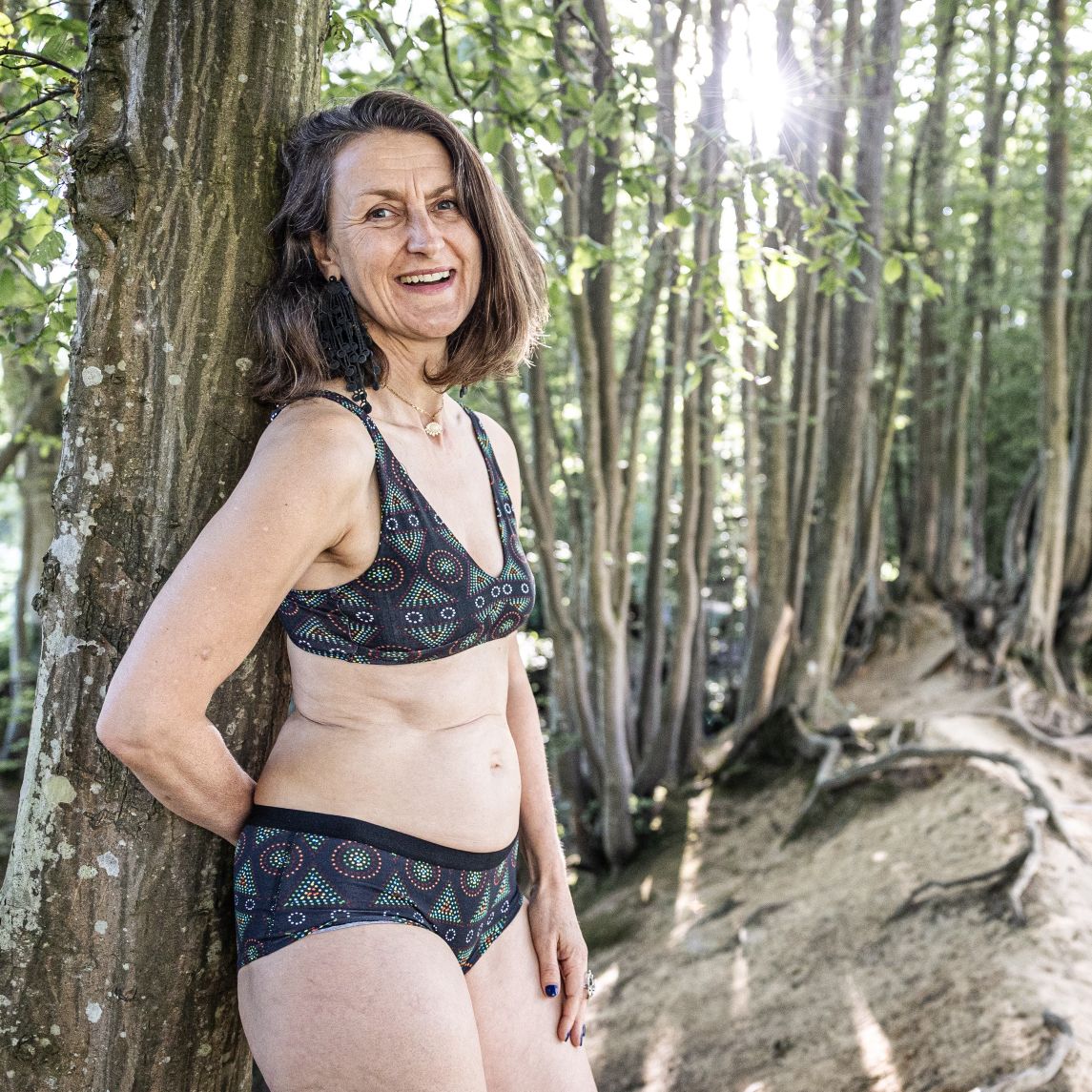 A photo of a model wearing the YOU Underwear bralette and matching boyshorts in the black mara print. She is standing side on to the camera and leaning against a tree. She is smiling and looking directly at the camera. Behind her, there is a woody and green forest.
