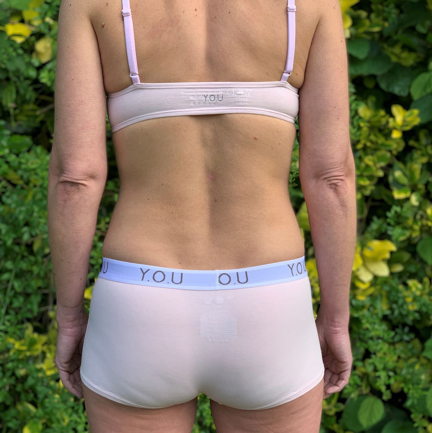 A photo of a model wearing the YOU Underwear bralette and boyshorts in light pink. She is facing away from the camera and there a green bush in front of her.