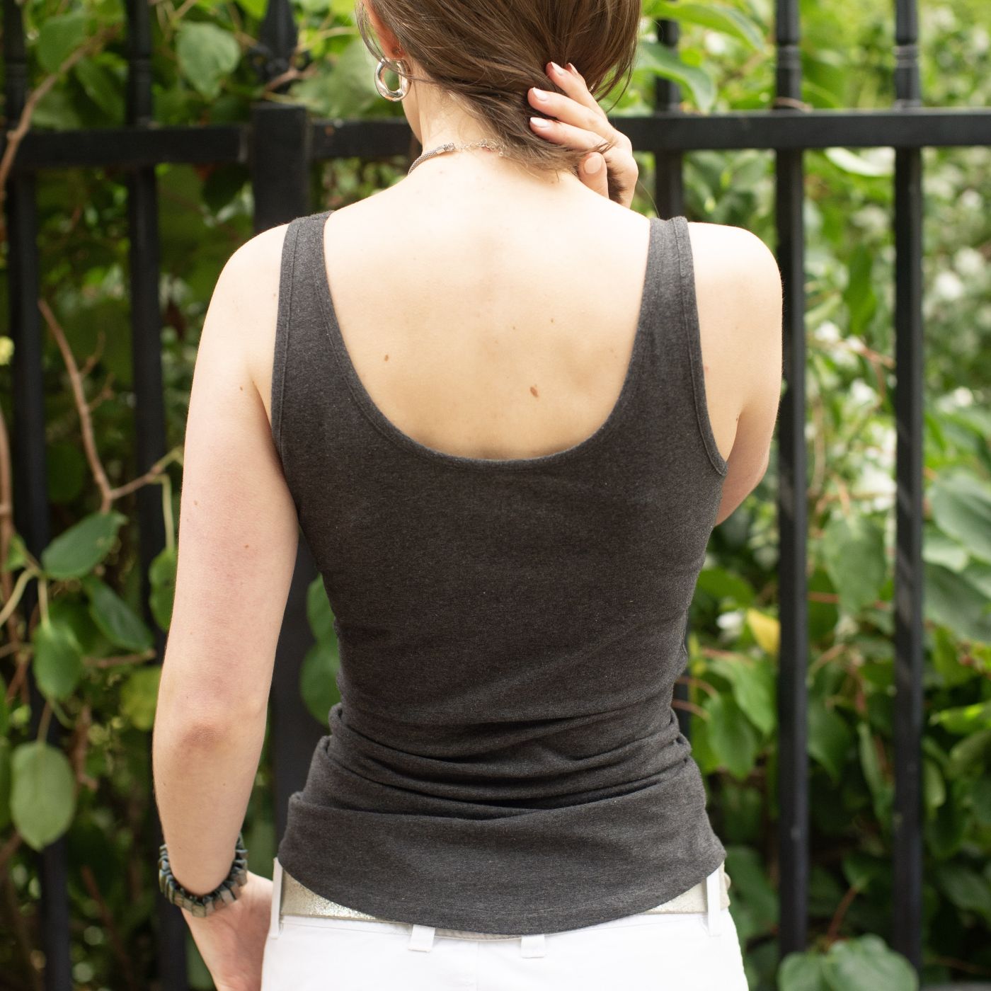 A photo of a model wearing the YOU Underwear vest in charcoal grey. They are facing away the camera, with their left hand down at their sides and the other holding her hair out of the way. The background is a black fence and a green bush.