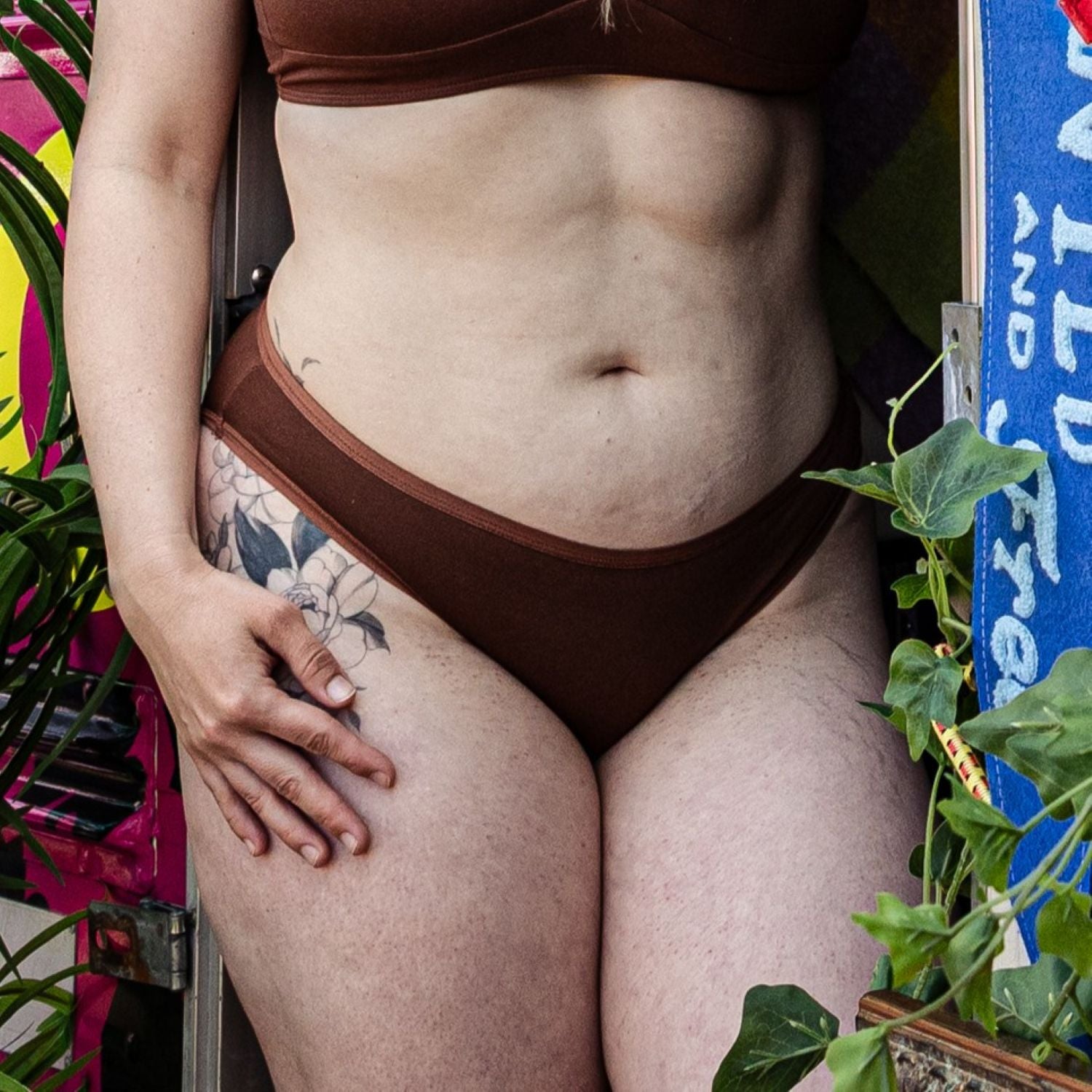 A photo of a model wearing the YOU Underwear low rise bikini knickers in chestnut. The photo is zoomed in so we can only see from the model's upper-stomach to their lower-thigh. She has her right hand on her right thigh, near a large flower tattoo. There is a green plant in the foreground of the photo.