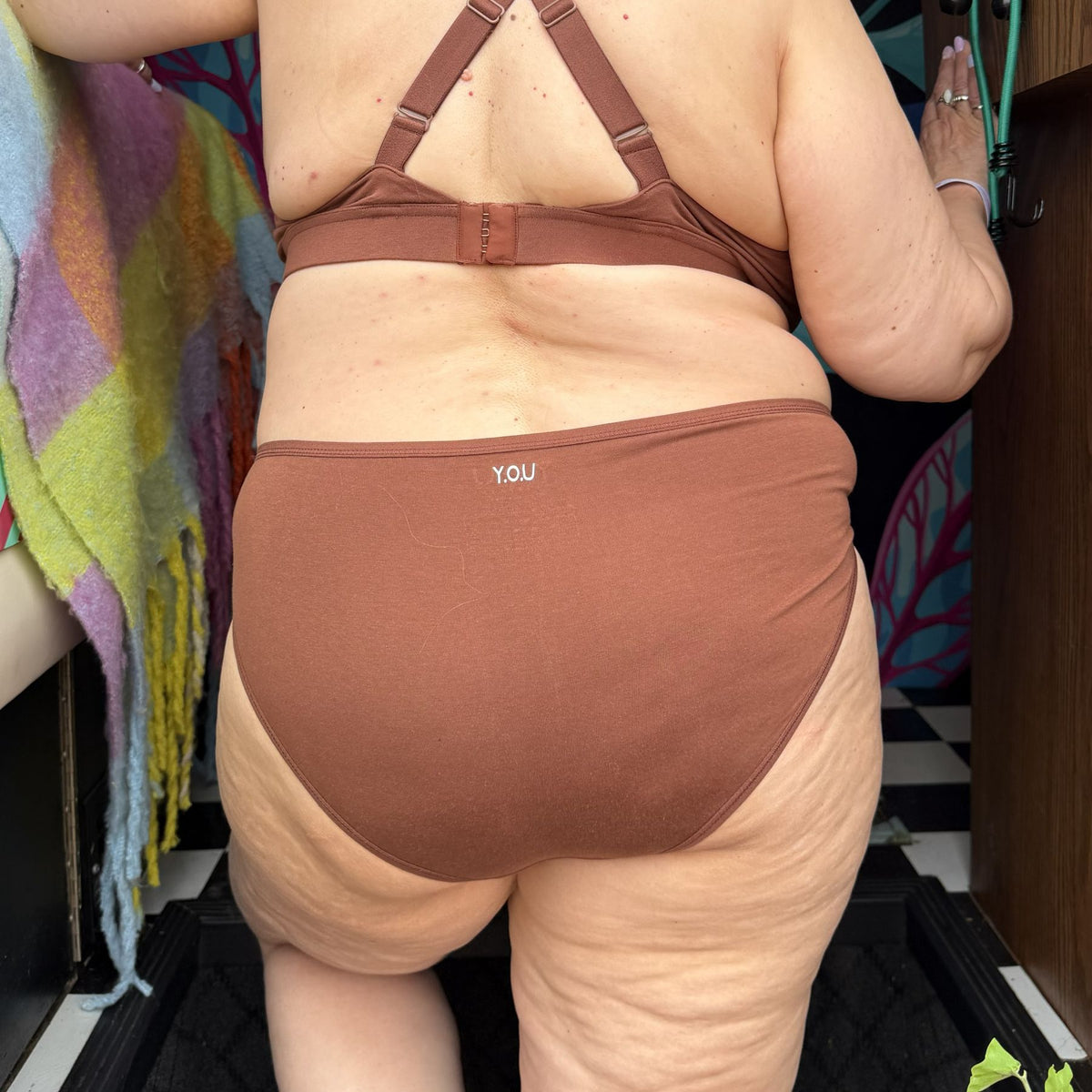 A photo of a model wearing the YOU Underwear mid-rise bikini knickers in the colour chestnut. The photo is zoomed in so we can see from the model's shoulder blades to their knees They are facing away from the camera. The background is dark but there is a colourful green, purple, blue and orange checked blanket to the left.