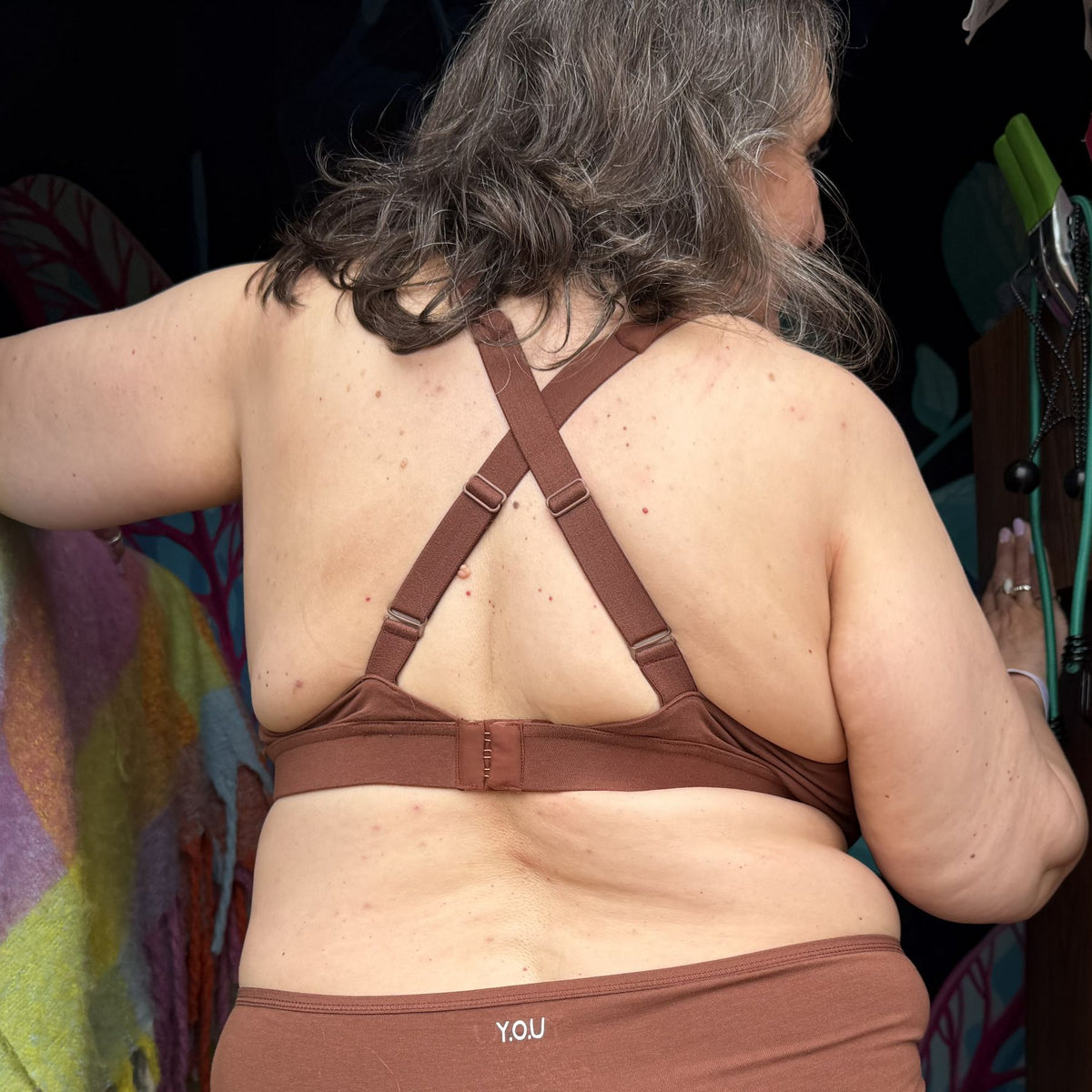 A photo of a model wearing the YOU Underwear non-wired bra and knickers in the colour chestnut. The photo is zoomed in so we can see from the model's shoulder's to their hips. They are facing away from the camera, showing off the adjustable straps which are crossed to show a different style of wear. The background is dark but there is a colourful green checked blanket to the left of the model.