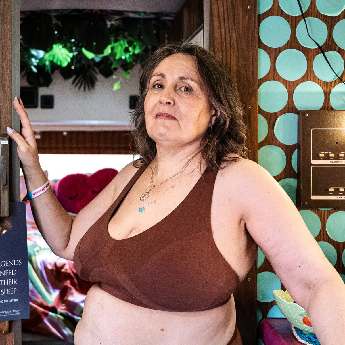 A photo of a model wearing the YOU Underwear non wired bra in chestnut. She is standing within a doorway with a hand on the frame. To her right there is a wall with bright blue polka dots. The photo is zoomed so that we can see to the model's waist. In the background there is some green foliage. 