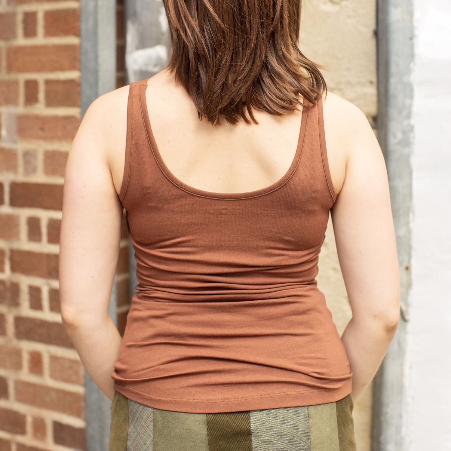 A model wearing the YOU Underwear chestnut vest She is standing in front of red brick wall, facing away from the camera with her hands in front of her. The photo is zoomed in so we can see from the model's shoulders to her hips.