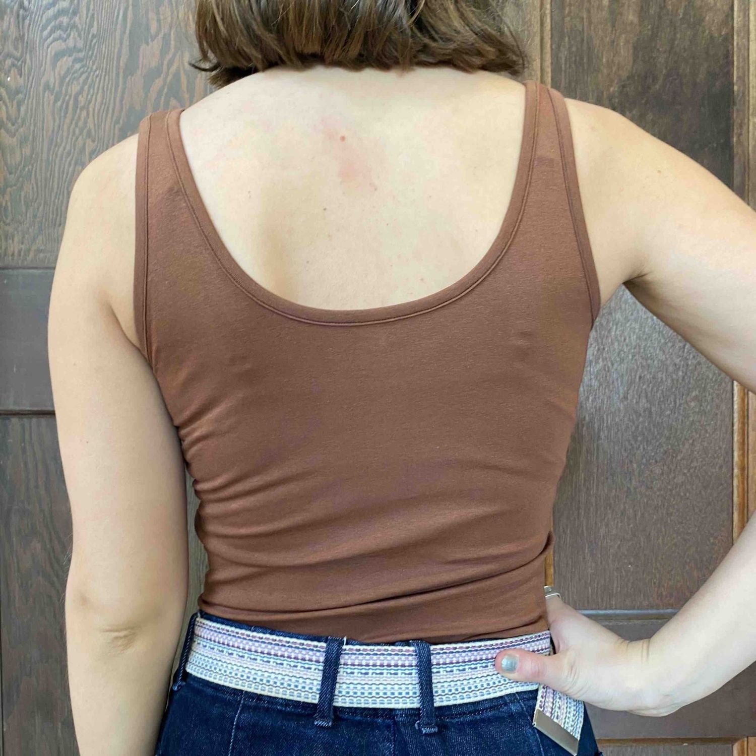 A photo of a model wearing the YOU Underwear vest top in chestnut. She is facing away the camera, with her right hand on her hip. The photo is zoomed in so we can see from the model's shoulders to their hips.