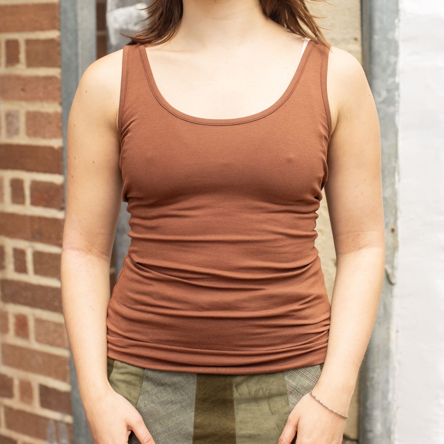 A model wearing the YOU Underwear chestnut vest She is standing in front of red brick wall, facing the camera with her hands in front of her. The photo is zoomed in so we can see from the model's shoulders to her hips.