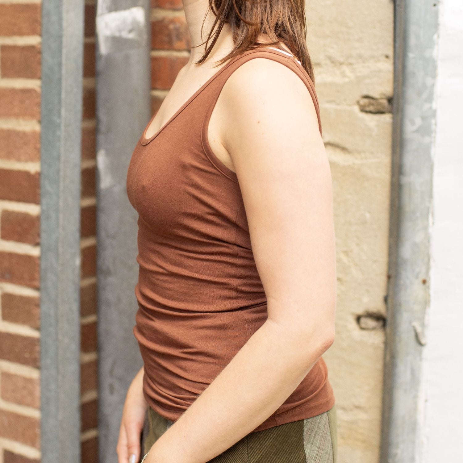 A model wearing the YOU Underwear chestnut vest She is standing in front of red brick wall, standing side on to the camera with her hands in front of her. The photo is zoomed in so we can see from the model's shoulders to her hips.