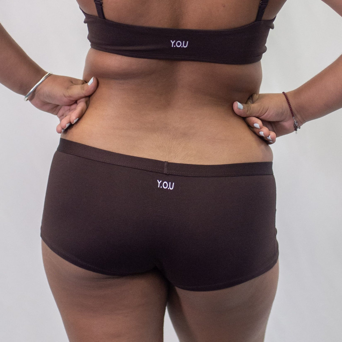 A photo of a model wearing the YOU Underwear cocoa boyshorts. They are standing facing away from the camera with both their hands on their hips. The photo is zoomed in so we can see from the model's shoulder blades to their upper-thigh. There is a white background.