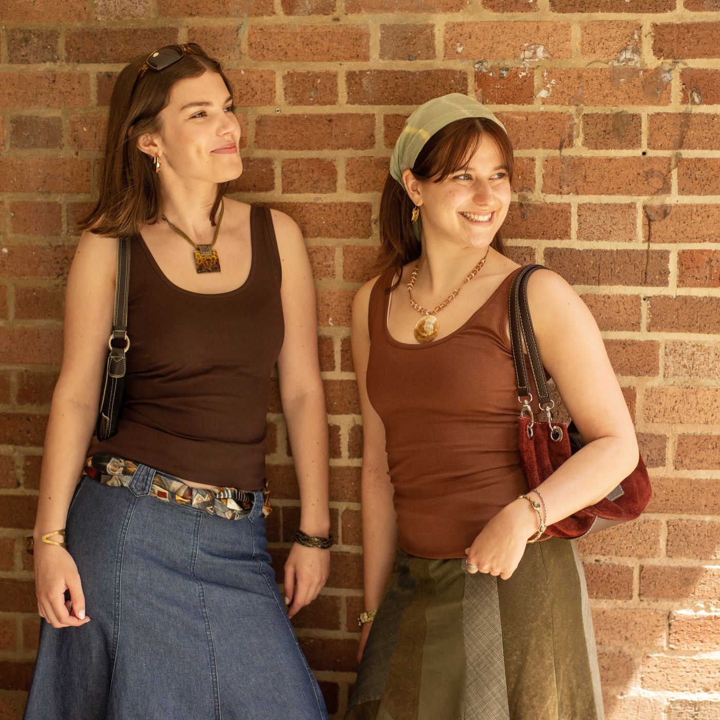 A picture of two women leaning against a red brick wall. One is wearing the YOU Underwear cocoa vest and the other is wearing the vest in chestnut. They have accessories such as handbags, sunglasses and head scarves. They are smiling looking out to their left.