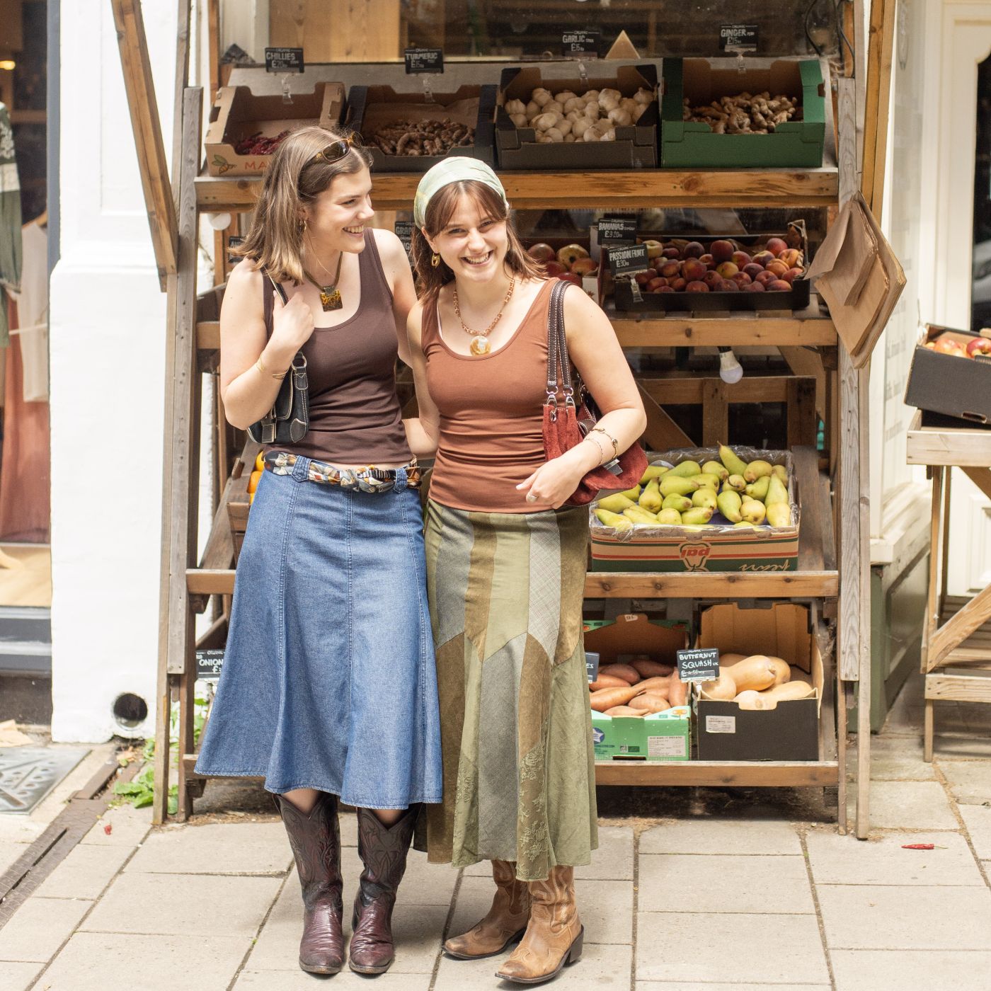 A picture of two women standing in front of a fruit and veg stall. One model is wearing the YOU Underwear cocoa vest paired with a denim skirt an cowboy boots and the other is wearing the YOU Underwear chestnut vest paired with a green skirt and cowboy boots. They are facing the camera, smiling and laughing with each other.