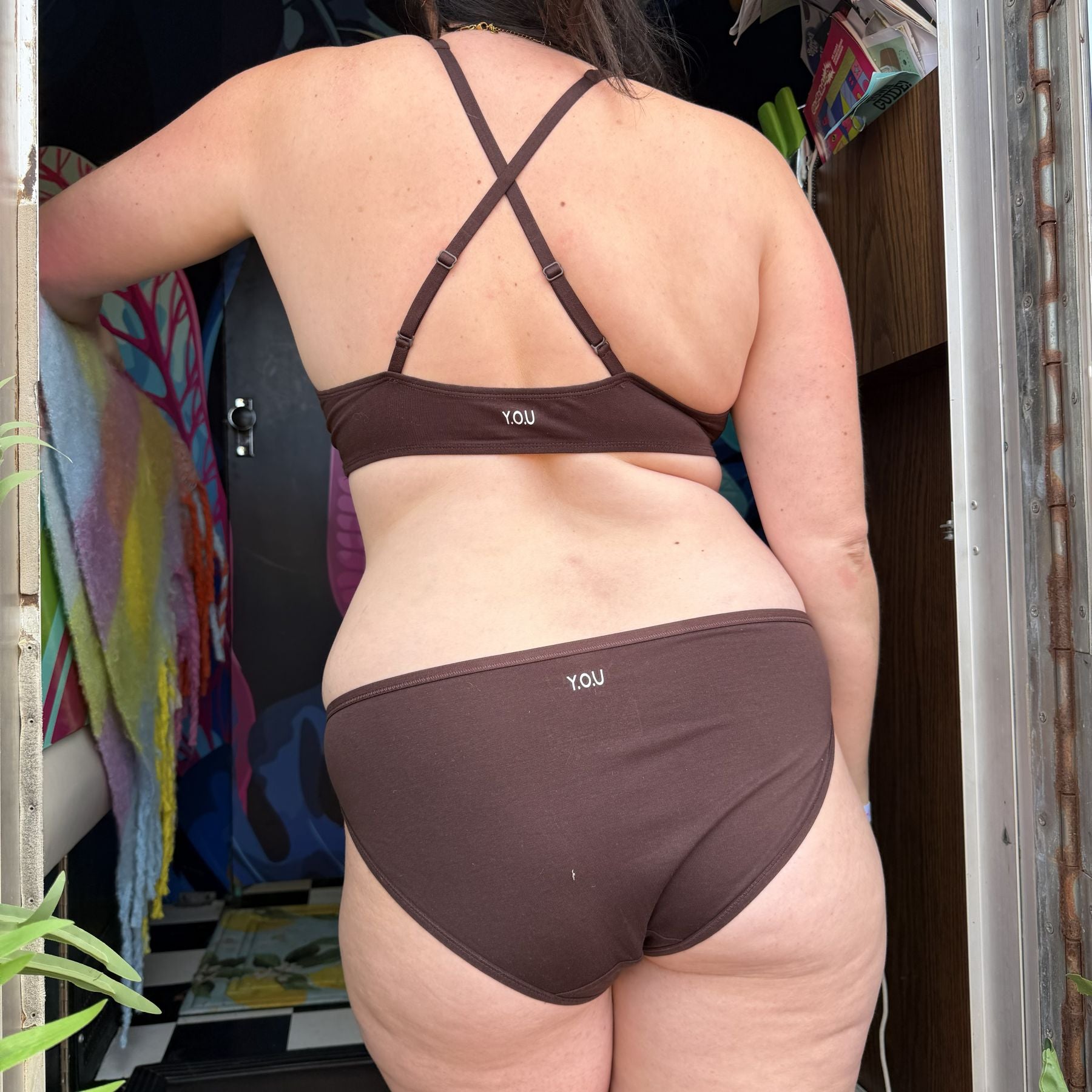 Close up image of model wearing YOU Underwear Bralette and Low Rise Bikini Matching Set in Cocoa. The model stands in a doorway, facing the camera. The bralette straps are crossed to show they are adjustable. The image shows from below the chin to the upper thigh