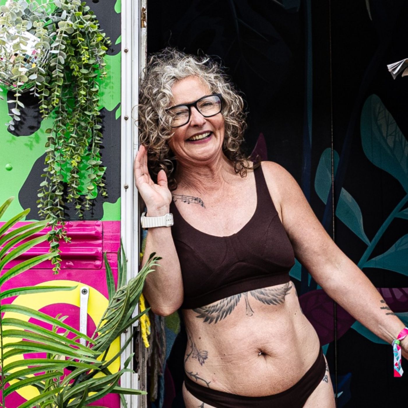 A photo of a model wearing the YOU Underwear non-wired bra and knickers in cocoa. She is standing, facing the camera and leaning against the doorway of a bright green leopard print bus. There is some green foliage to her left.