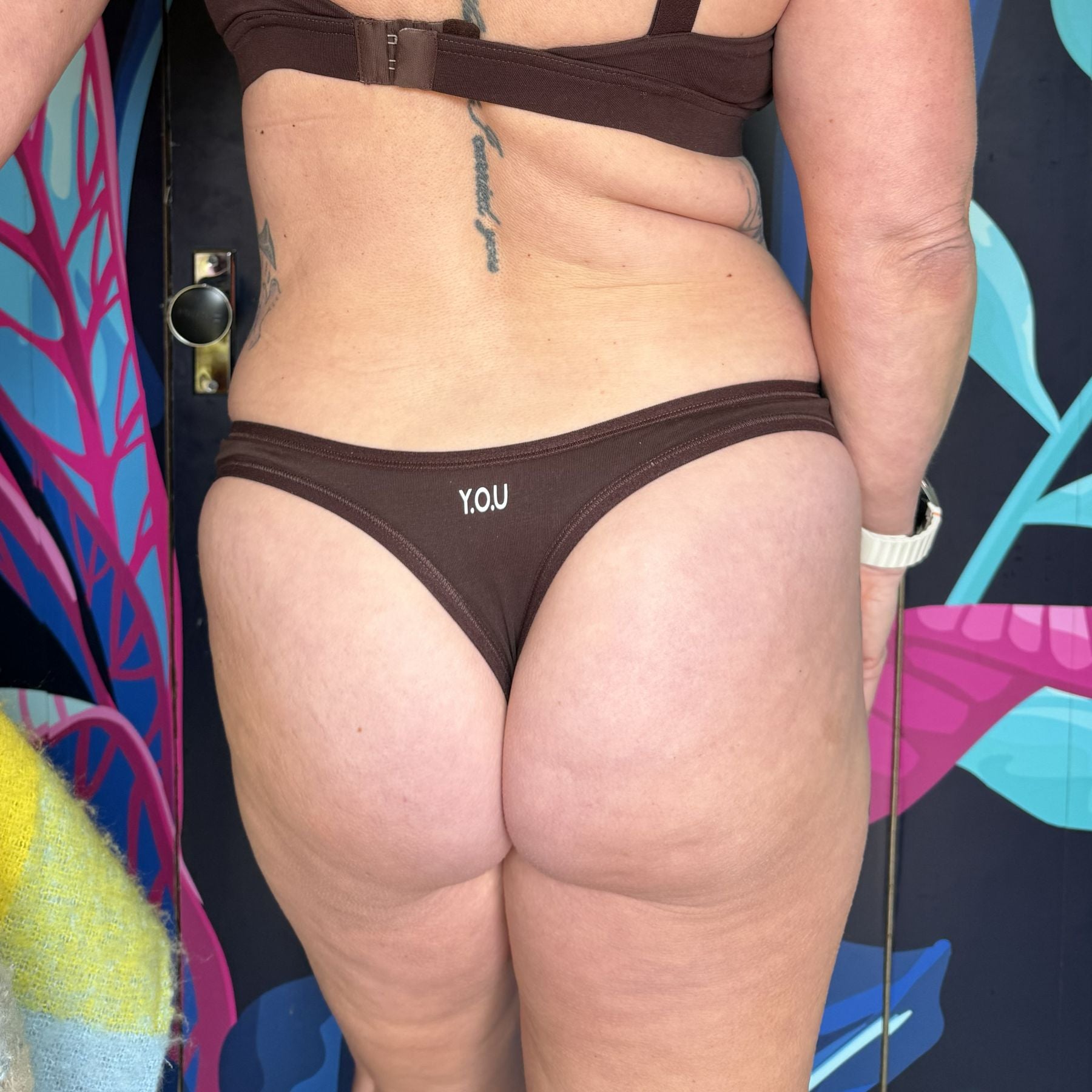 A photo of a model wearing the YOU Underwear thong in the colour cocoa. The photo is zoomed in so we can see from the model's shoulder blades to their upper-thigh. The model is facing away from the camera and has a tattoo running down her spine. The background is a dark blue wall with bright blue and pink leaves painted onto it.
