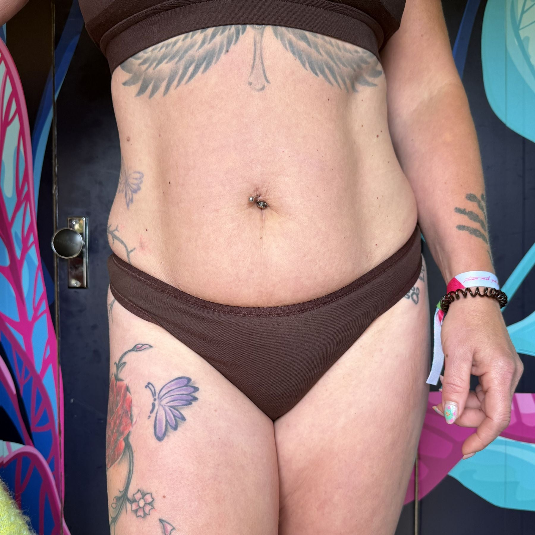 A photo of a model wearing the YOU Underwear thong in the colour cocoa. The photo is zoomed in so we can see from the model's stomach to their upper-thigh. The model is facing the camera and has various tattoos over her body. Behind her is a dark blue wall with bright blue and pink leaves painted onto it.