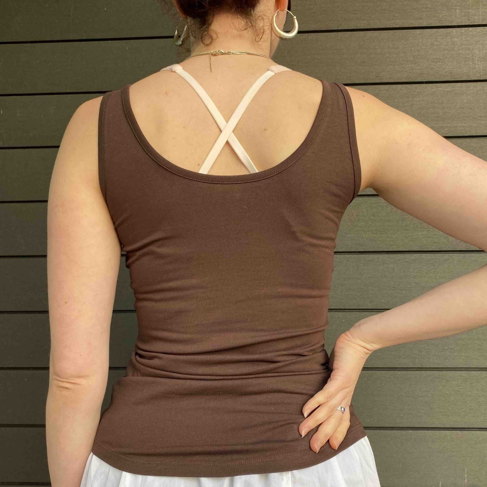 A close up picture of a model wearing the YOU Underwear vest in cocoa. She is standing facing away the camera, with her right hand on her hip. There is a wooden paneled wall in the background. The photo is zoomed in so we can see from the model's neck to their hips.