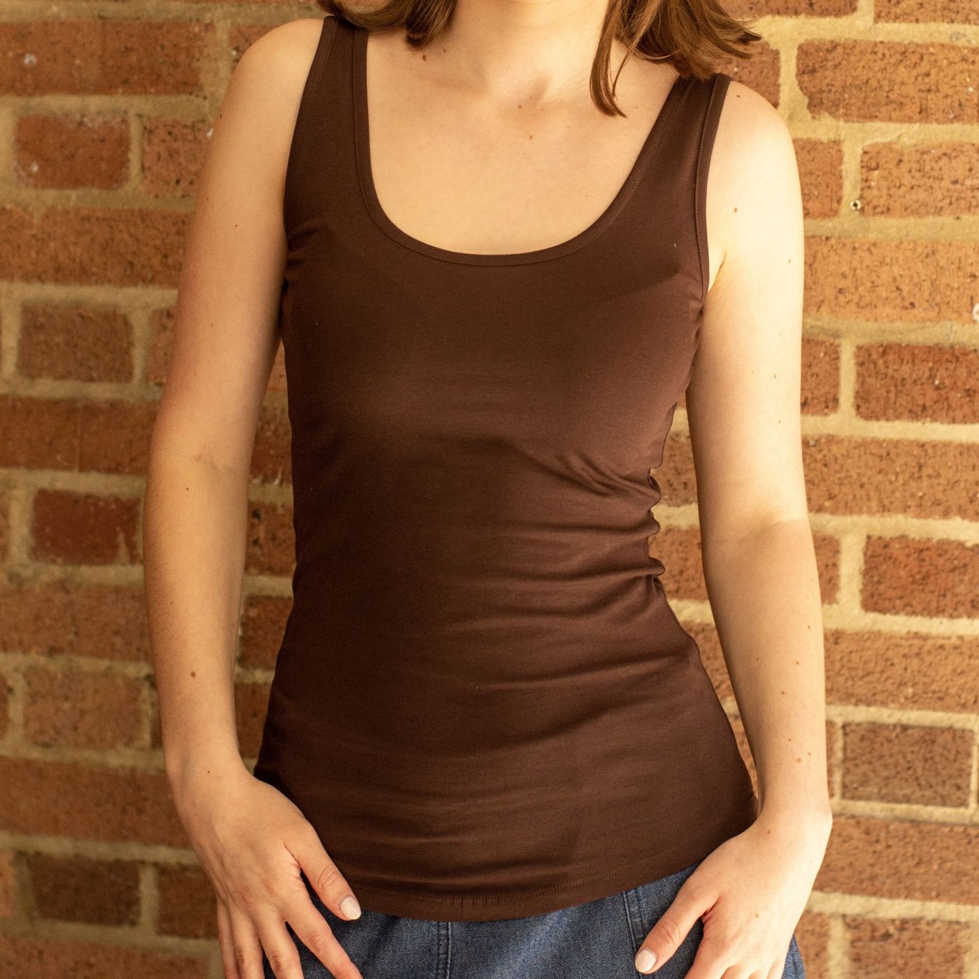 A product focused photo of a model wearing the YOU Underwear vest in cocoa. She is standing facing the camera, with her hands in front of her. The photo is zoomed in so we can see from her shoulders to her hips. There is a red brick wall behind her.