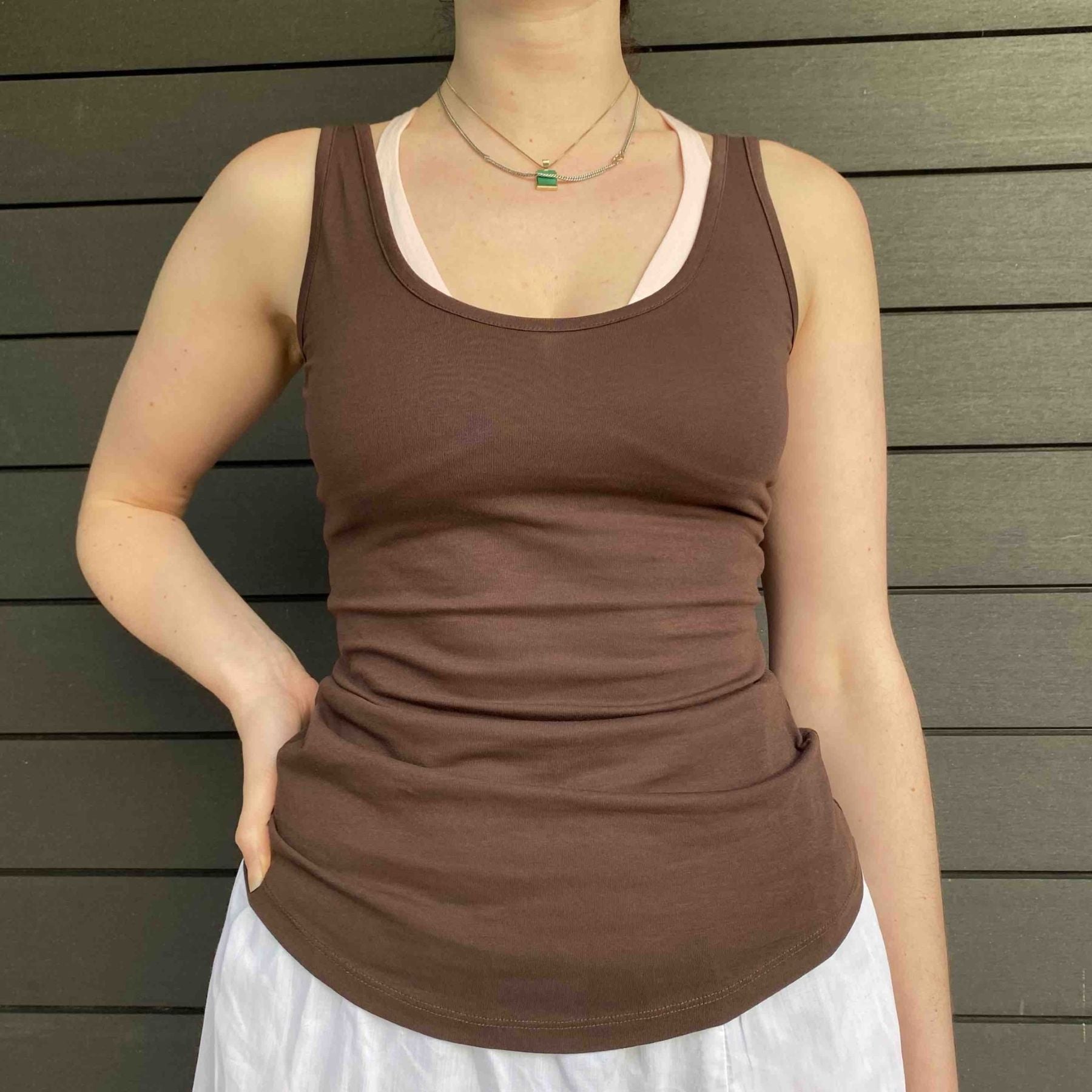 A close up picture of a model wearing the YOU Underwear vest in cocoa. She is standing facing the camera, with her right hand on her hip. There is a wooden paneled wall in the background. The photo is zoomed in so we can see from the model's neck to their hips.