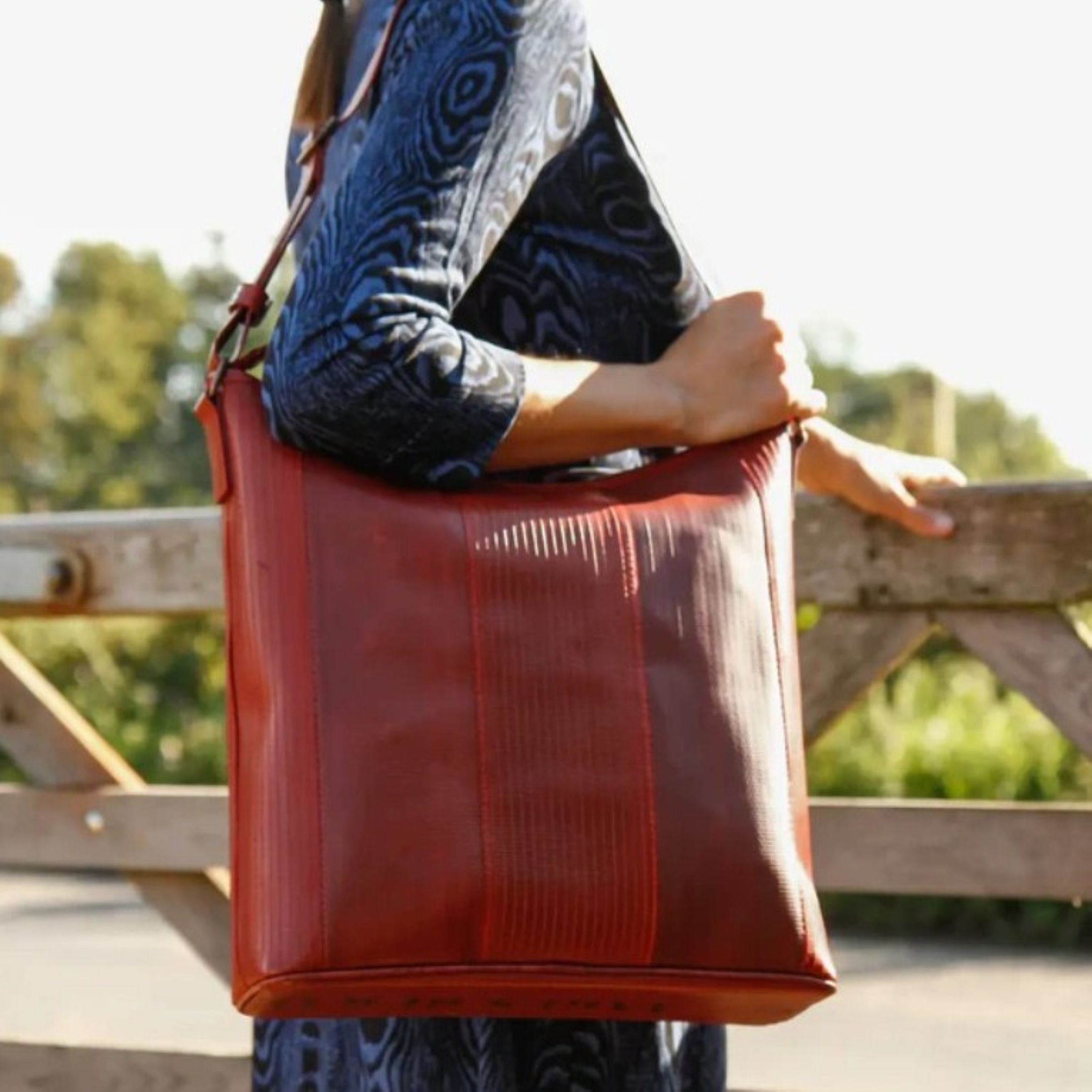 A close up photo of a woman holding the Elvis & Kresse red Tooley Tote bag over her right shoulder, leaning on a gate and looking towards green trees