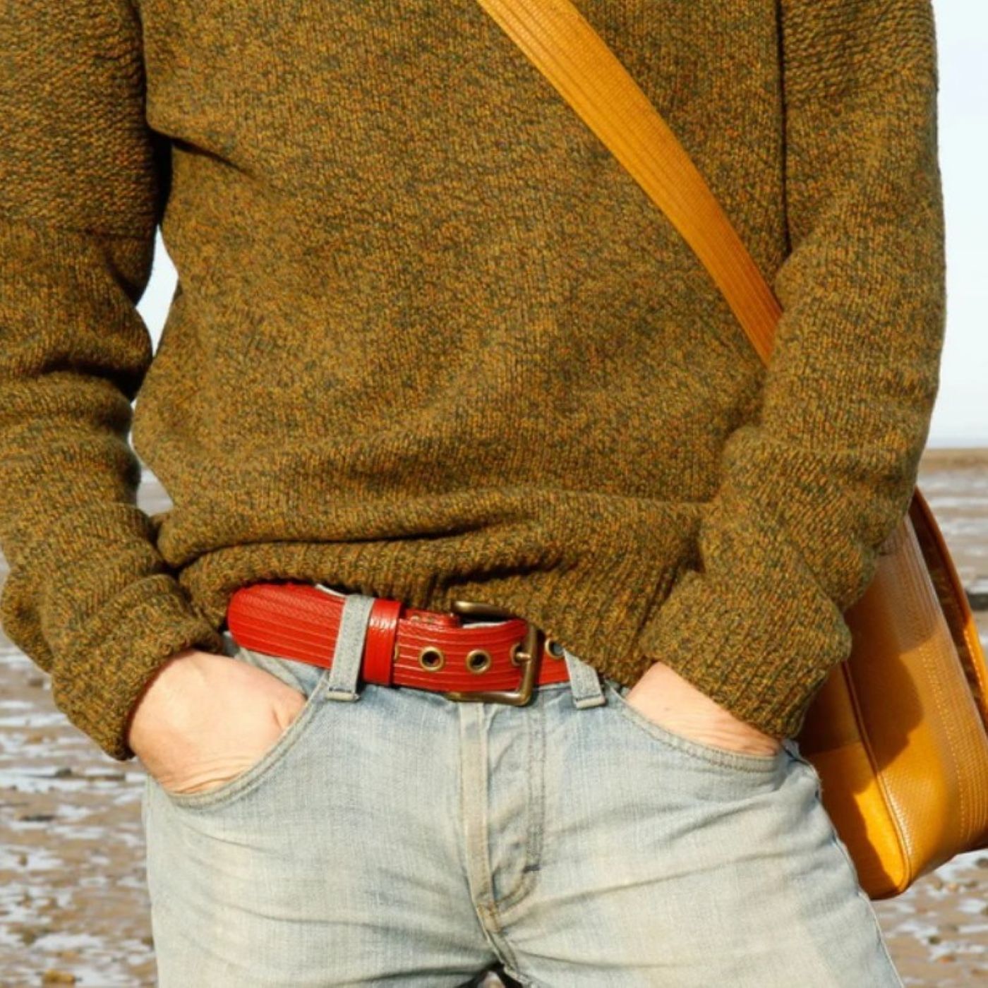 A close up photo of a model wearing the Elvis & Kresse red west end belt paired with a yellow and green knitted jumper, pale blue jeans and a yellow cross body bag. The picture is taken with the backdrop of a beach.