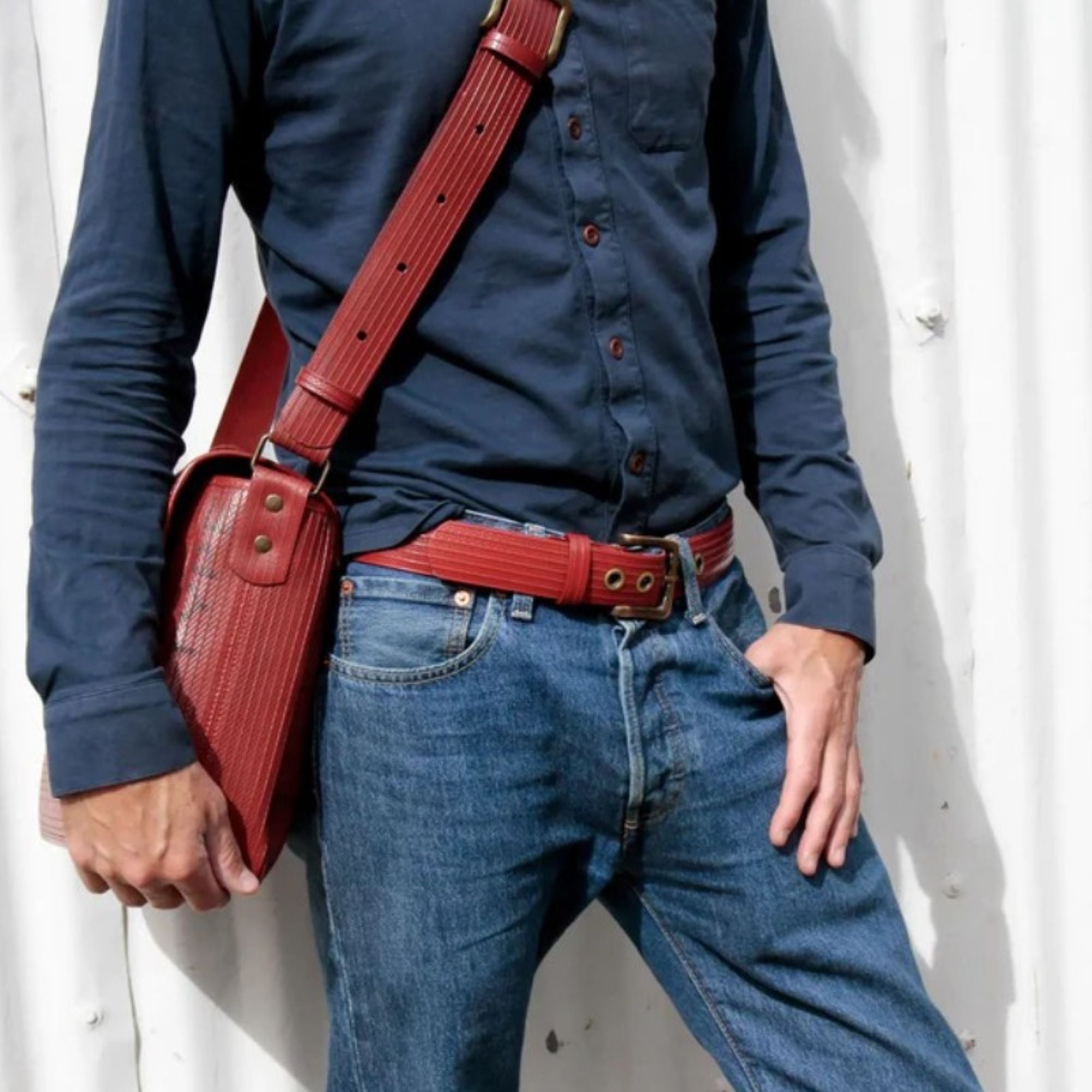 A close up photo of a model wearing the Elvis & Kresse red west end belt paired with dark blue jeans, a dark blue shirt and the Elvis & Kresse red cross body bag. The model is standing in front of a white corrugated metal background.