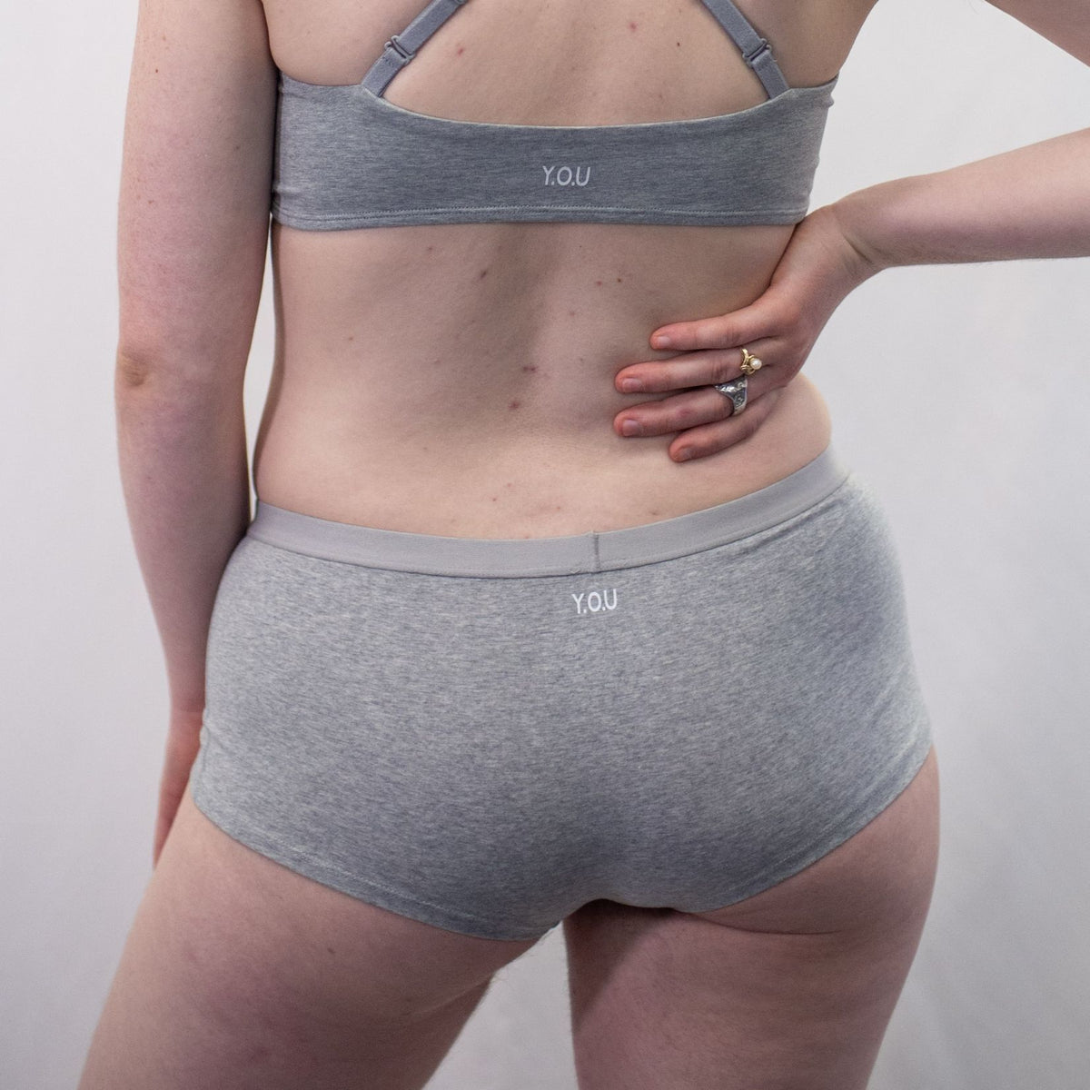 A photo of a model wearing the YOU Underwear boyshorts in grey with the matching bralette. The model is standing facing away from the camera with their right hand on their hip and the other at their side. The photo is zoomed in so we can see from the model's shoulder blades to their upper-thigh. The background is white.