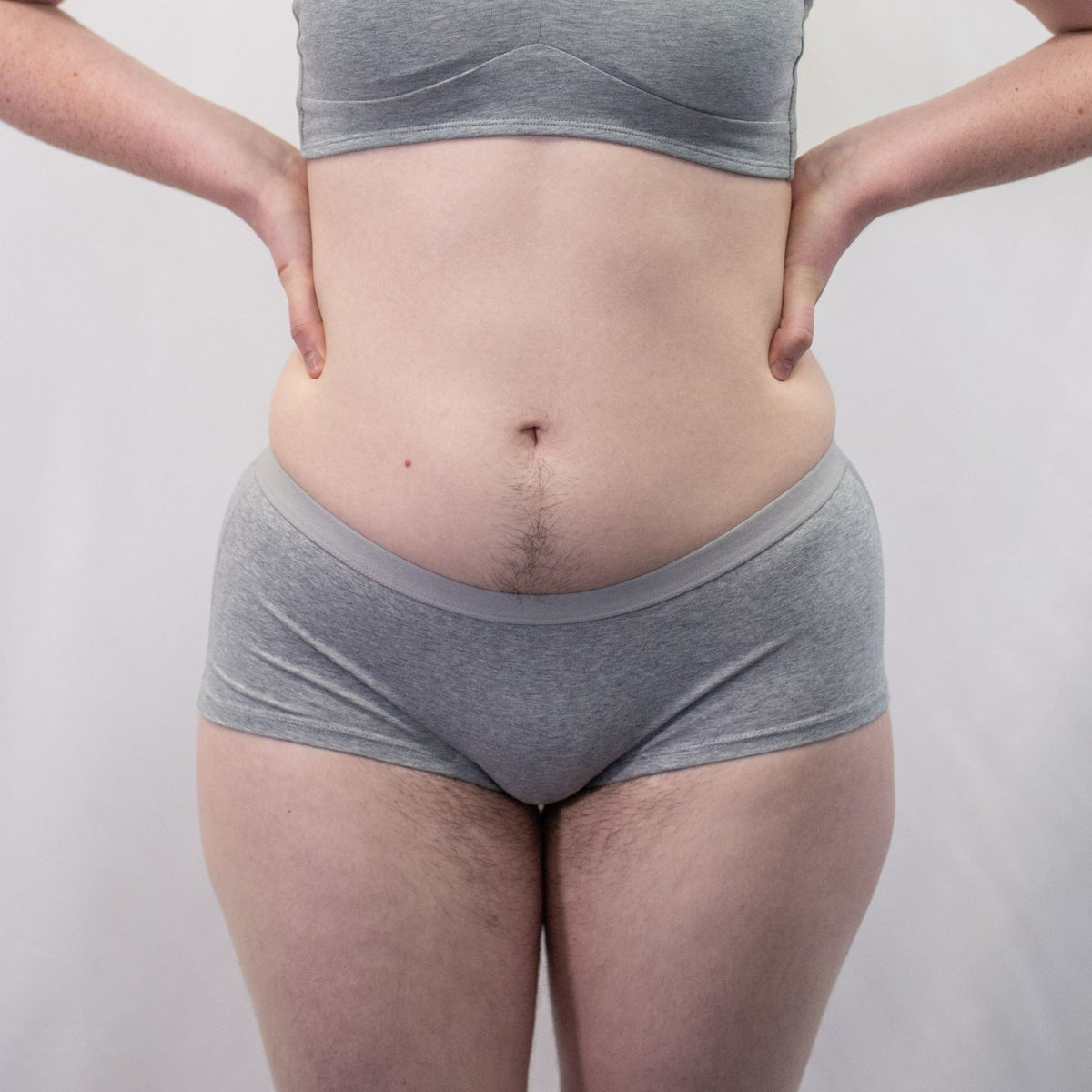 A photo of a model wearing the YOU Underwear boyshorts in grey with the matching bralette. The model is standing facing the camera with their hands on their hips. The photo is zoomed in so we can see from the model's stomach to their upper-thigh. The background is white.