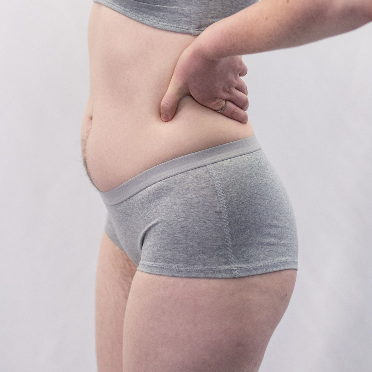 A photo of a model wearing the YOU Underwear boyshorts in grey with the matching bralette. The model is standing side on to the camera with their hands on their hips. The photo is zoomed in so we can see from the model's stomach to their upper-thigh. The background is white.