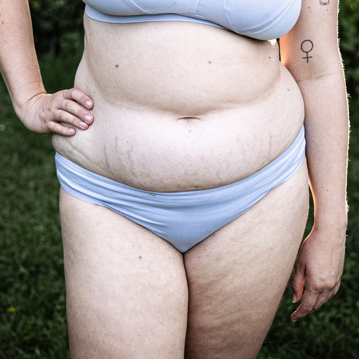 A photo of a model wearing the YOU Underwear low rise bikini knickers in light blue. The photo is zoomed in so we can see from the models chest to their lower-thigh. We can see that they have their right hand on their hip and there is lots of greenery in the background.
