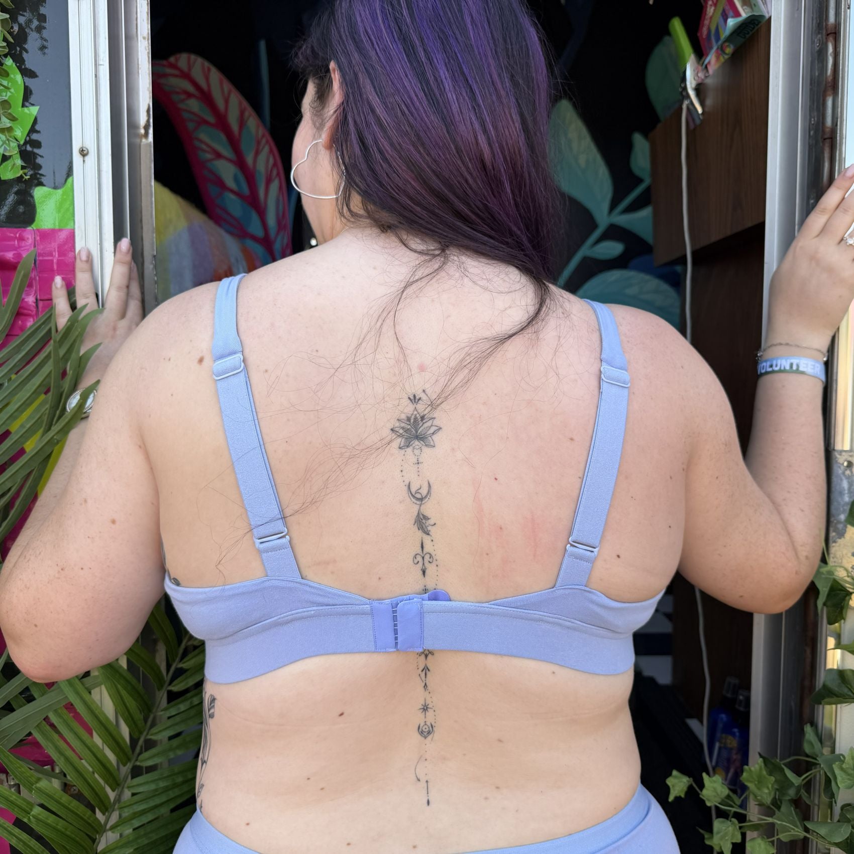 A photo of a model wearing the YOU Underwear non-wired bra in light blue. The photo is zoomed in so we can see from the model's head to her hips. She is facing away from the camera. There is some green foliage to the left of her and she appears to be standing in a doorway.