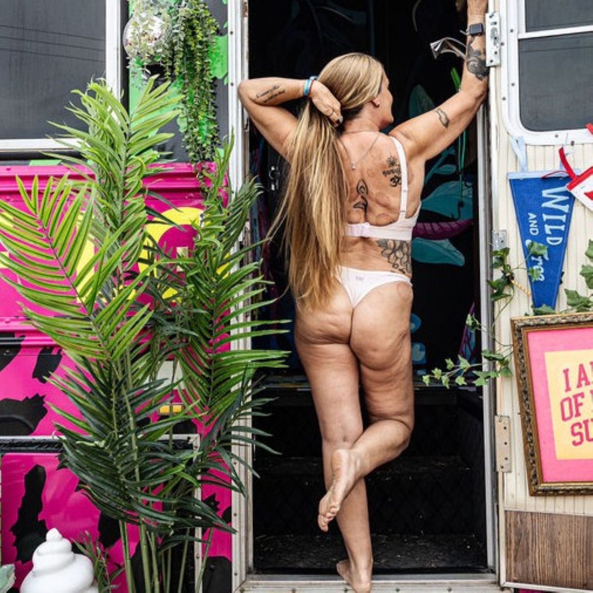 A photo of a model wearing the YOU Underwear light pink bra and thong. She is standing in the doorway of a camper-van, facing away from the camera. Her right arm is leaning against the door from and her left is pulling her long hair over her shoulder. There is a green plant in the left and the van is painted with a bright pink and black leopard print.