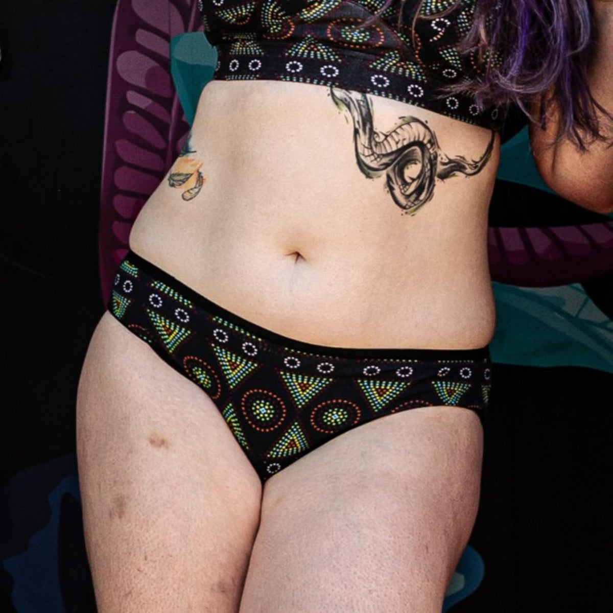 A photo of a model wearing the YOU Underwear low rise bikini knickers in black mara. The photo is zoomed in so we can see from the model's chest to their mid-thigh. 