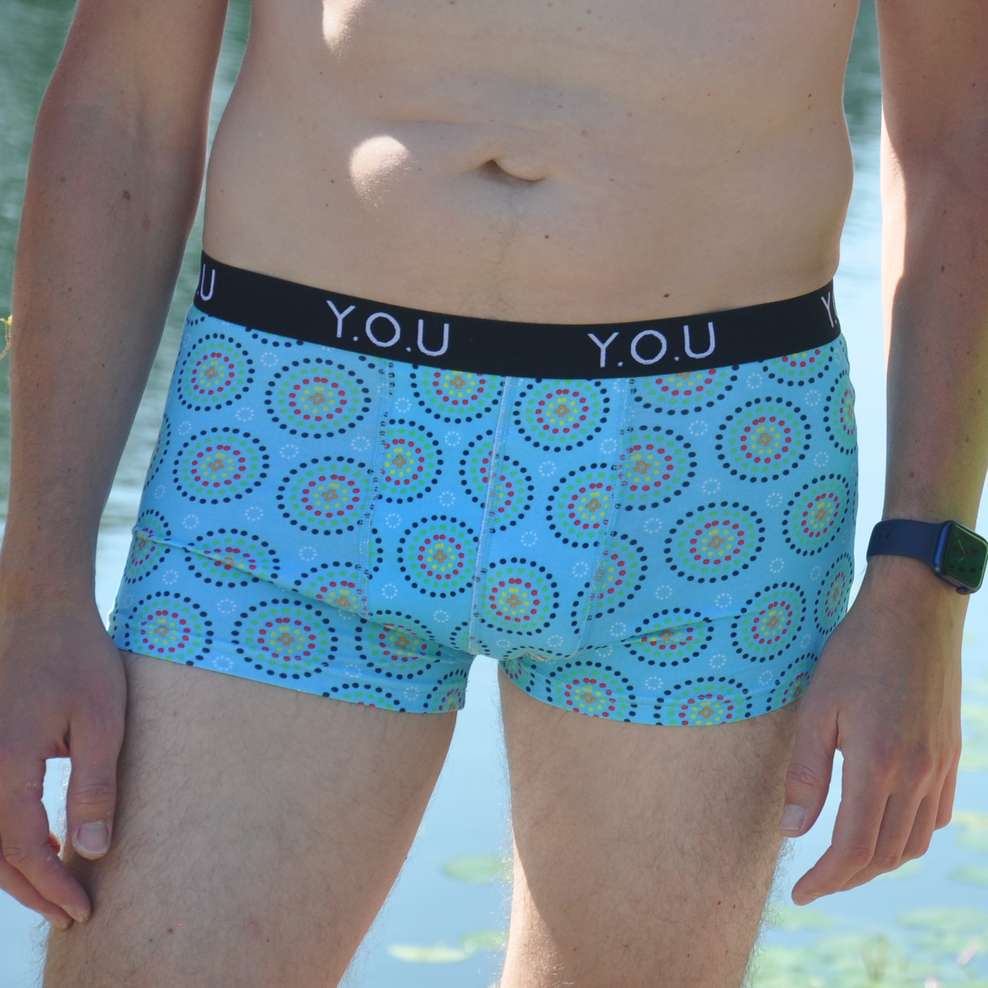 A person is standing by a body of water, wearing blue hipster trunks with a multicolored circular pattern. The waistband of the trunks has the text "Y.O.U." printed on it. The person is also wearing a dark wristwatch. Their upper body and legs are partly visible.