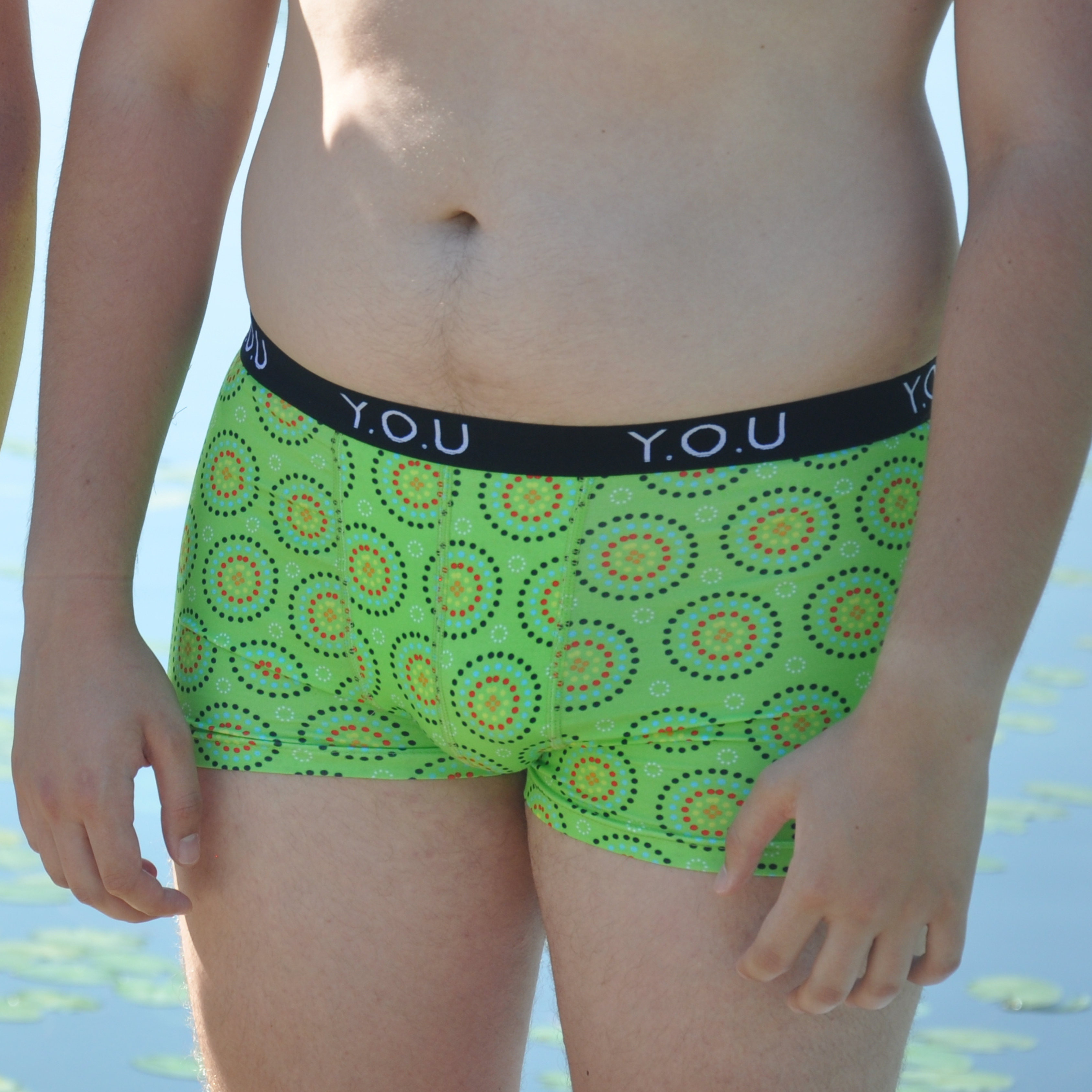 A person standing outdoors near water, wearing bright green men's hipster trunks with colorful circular prints and a black waistband labeled "Y.O.U". The person's face and surrounding environment are not visible.