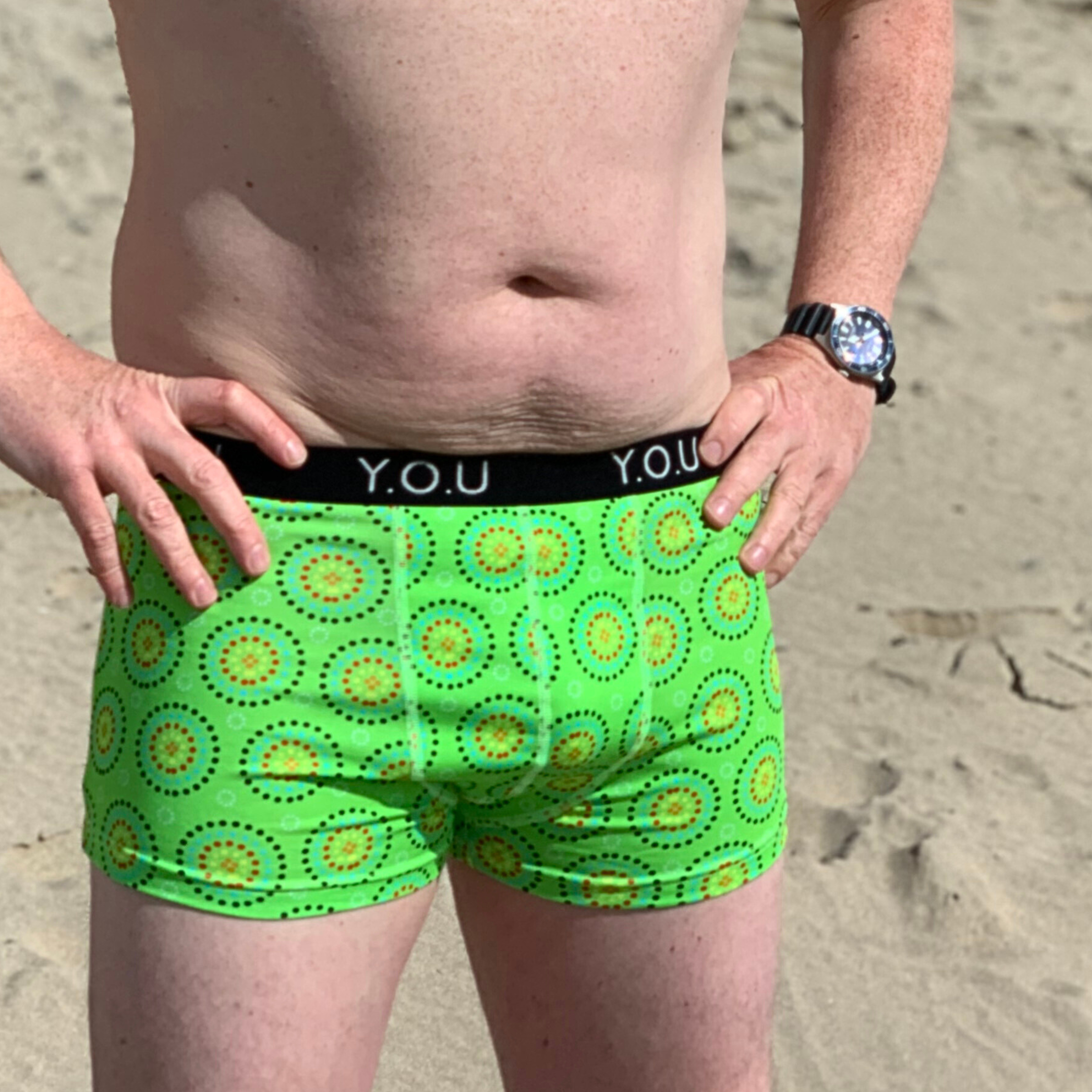A person standing on a sandy beach, wearing bright green shorts with a circular print and a waistband that reads "Y.O.U." They are shirtless and wearing a watch on their left wrist. Their hands are resting on their hips.