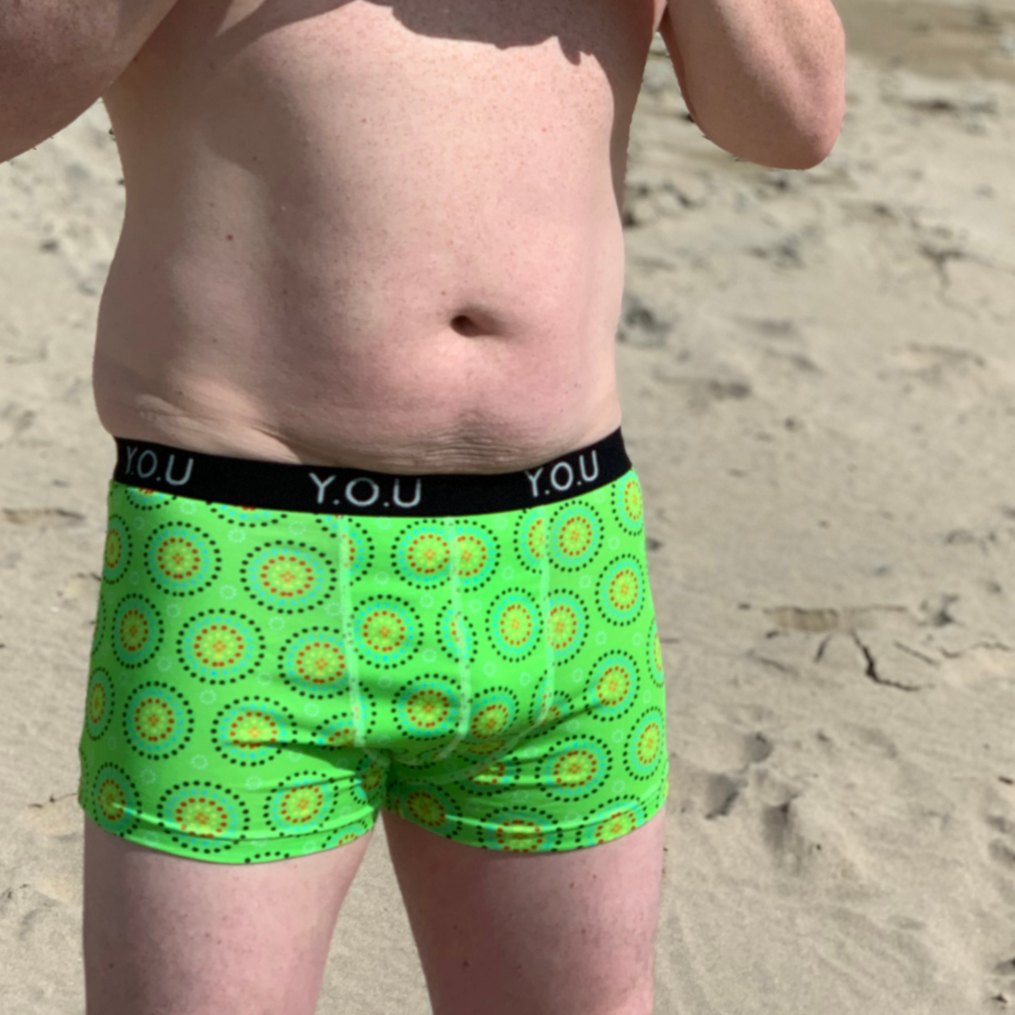 A person with a bare torso stands on a sandy beach. They are wearing bright green men’s hipster trunks adorned with yellow and red circular patterns, and a black waistband that has "Y.O.U" written on it repeatedly. The person's face is not visible.