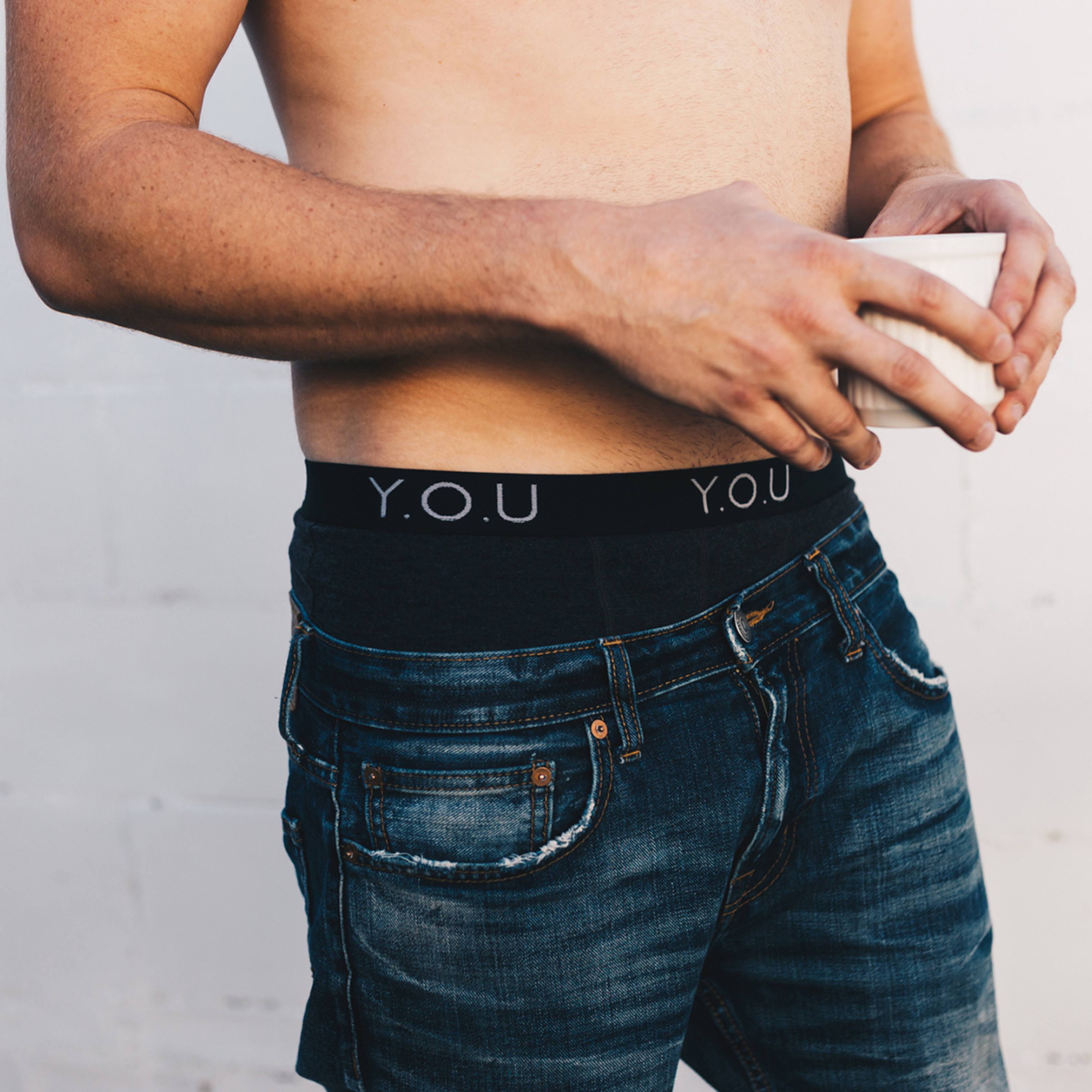 A shirtless person is holding a white ceramic cup with both hands, wearing dark blue jeans and black men's hipster trunks with the letters "Y.O.U" visible on the waistband. The background is a plain, pale-coloured wall.
