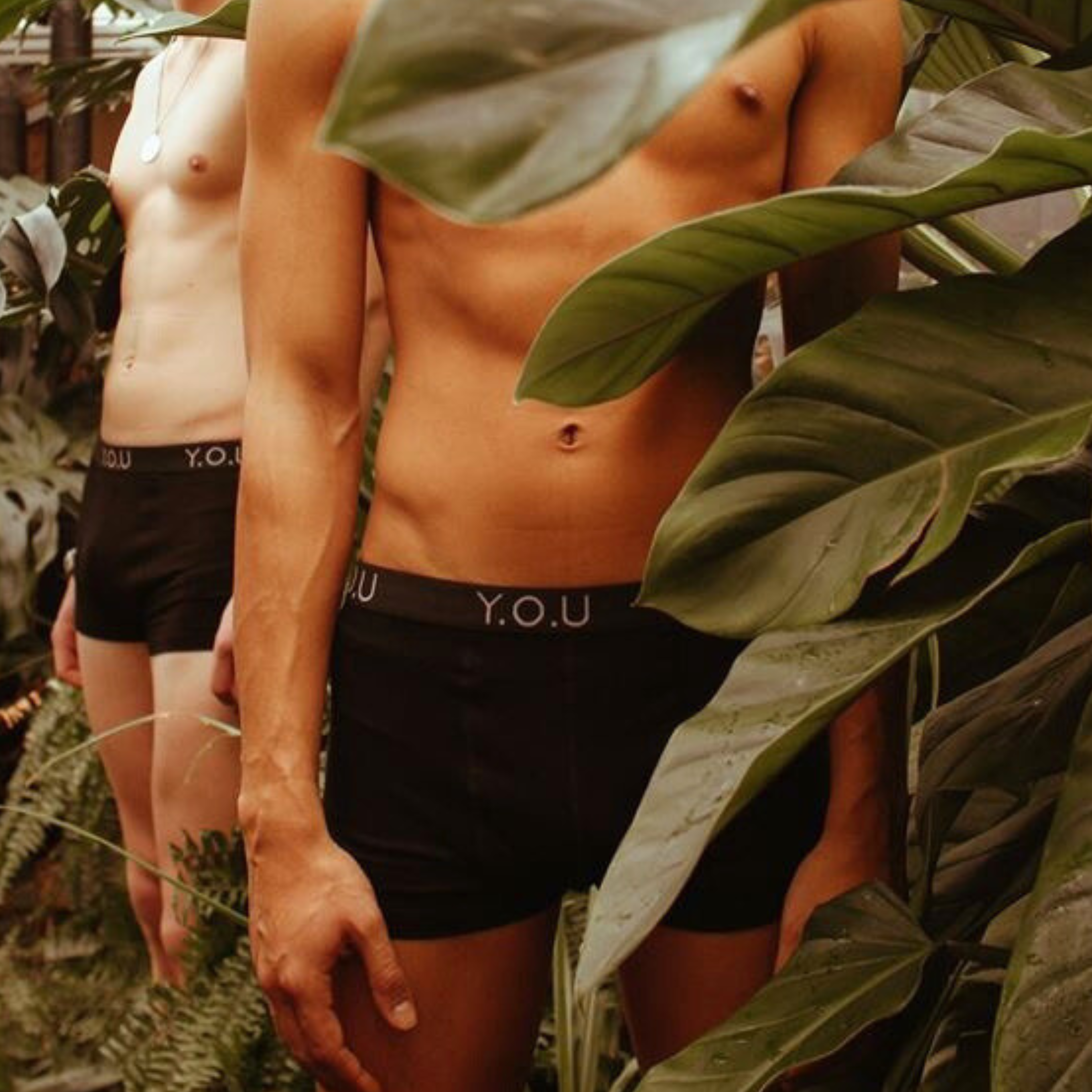 Two individuals wearing men's black hipster trunks are partially visible amidst lush green indoor plants. They are both shirtless, and the person in the foreground stands with arms relaxed at their sides. Their underwear has the initials "Y.O.U" on the waistband.
