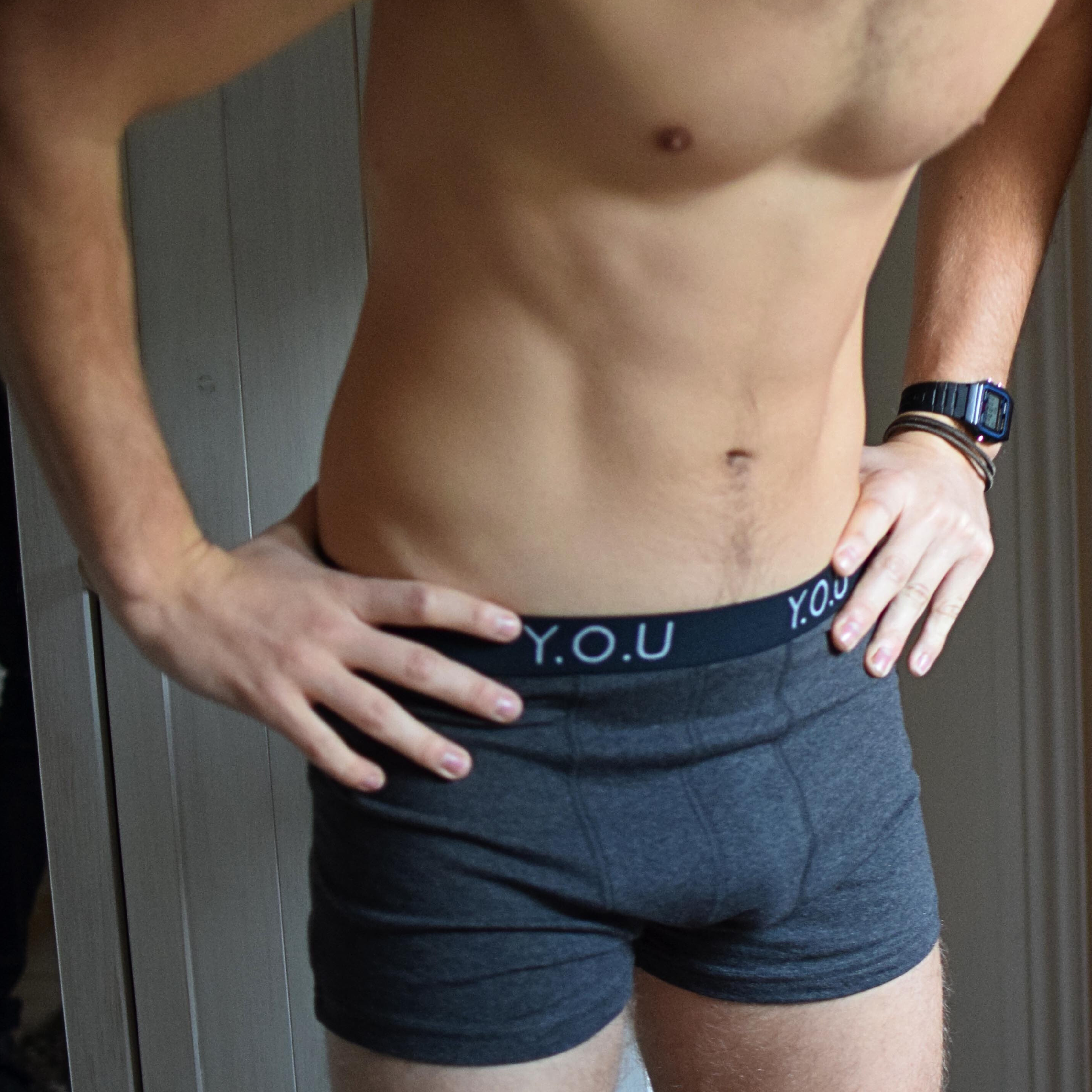 A shirtless person wearing dark grey organic cotton trunks with "Y.O.U" printed on the waistband stands with hands on their hips. They are also wearing a black wristwatch and a bracelet on their left wrist. The background features a wooden surface.