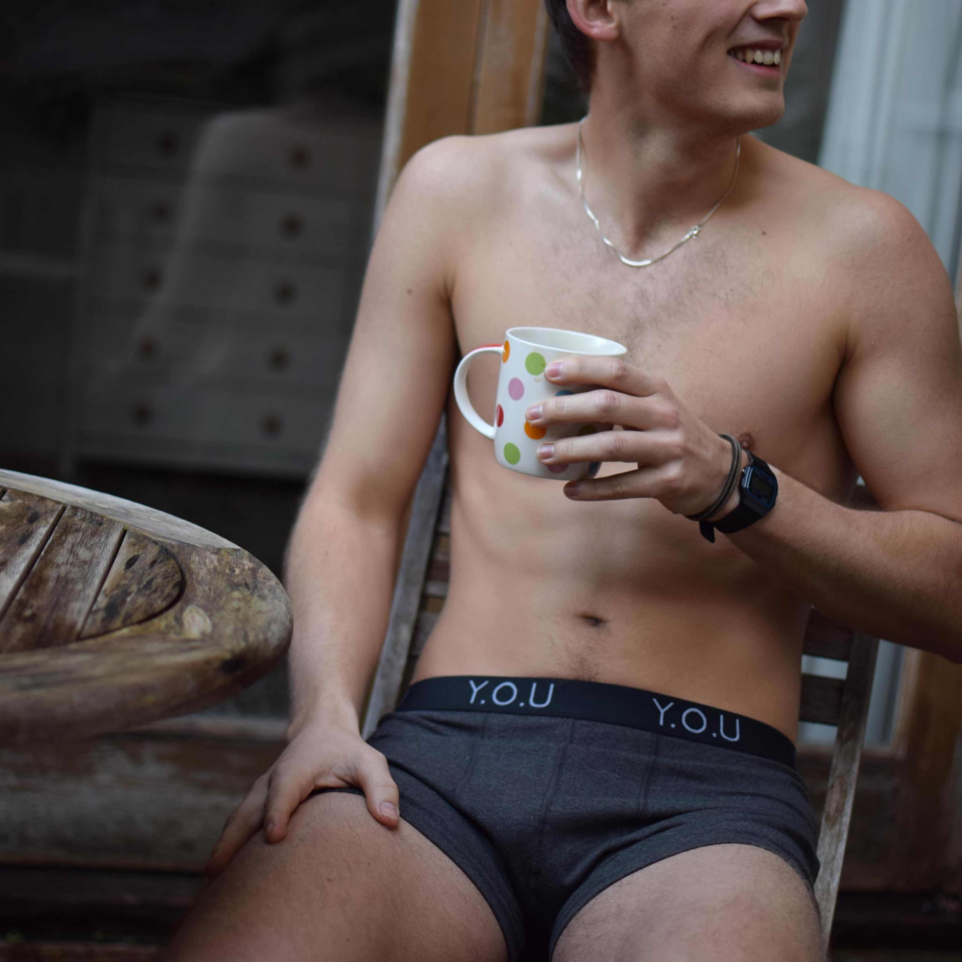 A shirtless person is sitting on a wooden chair, sporting grey men’s hipster trunks with "Y.O.U" on the waistband. They hold a polka-dotted coffee mug and smile while looking to the side. A silver necklace and wristwatch are visible.