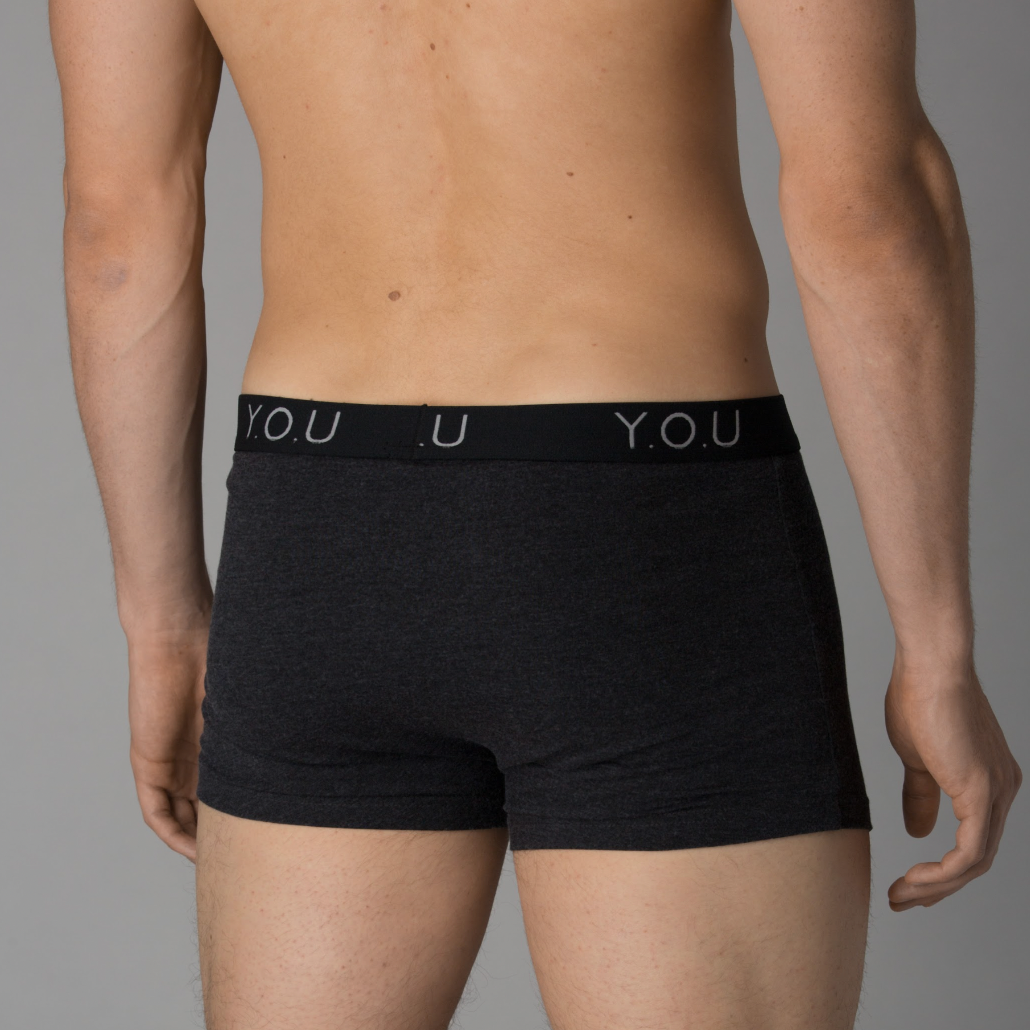 A person is standing with their back to the camera, modeling a pair of dark grey men's hipster trunks. The waistband of the hipster trunks is black with the letters "Y.O.U" in white. 