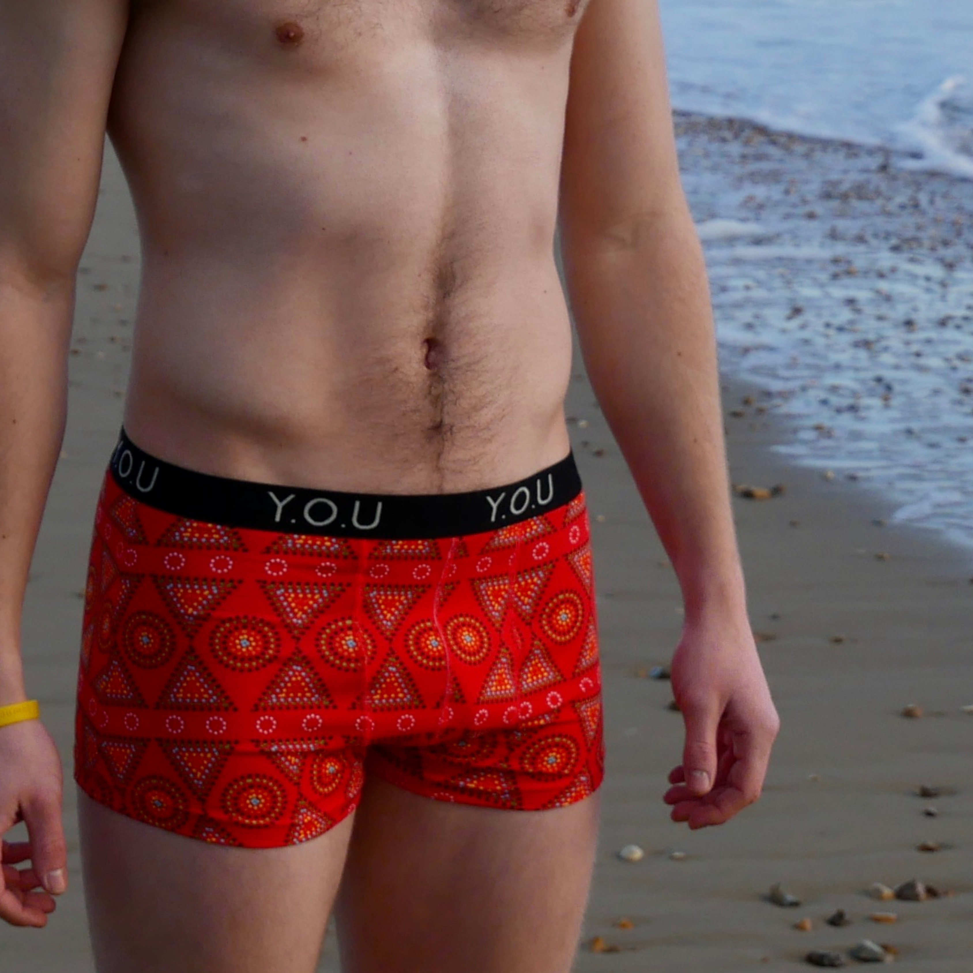 A person standing on the beach wearing red patterned men's hipster trunks with "Y.O.U" on the waistband. The sea and a sandy shoreline are visible in the background. The person has a yellow wristband on their right wrist.